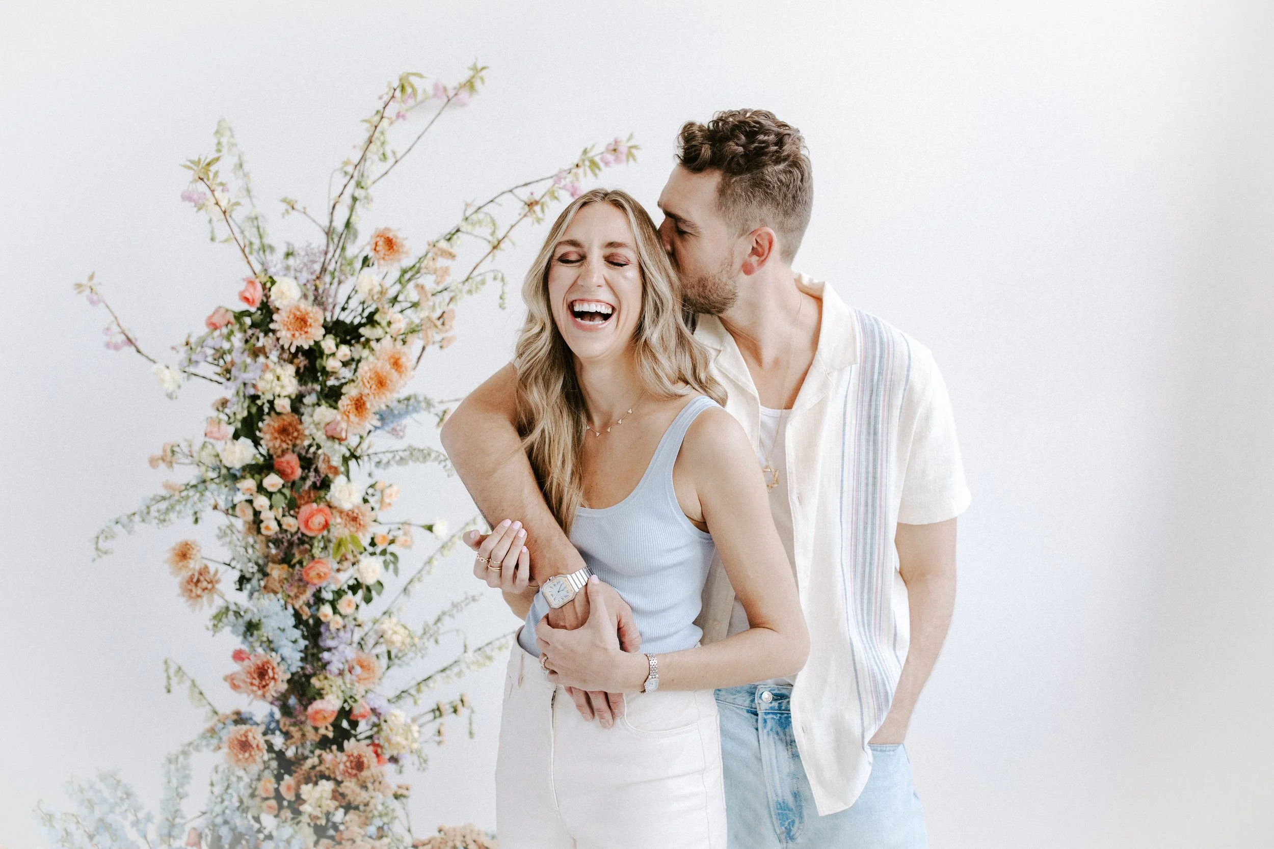 A joyful couple standsnext to a tall floral arrangement; the man kisses the woman on the temple while she smiles and laughs.