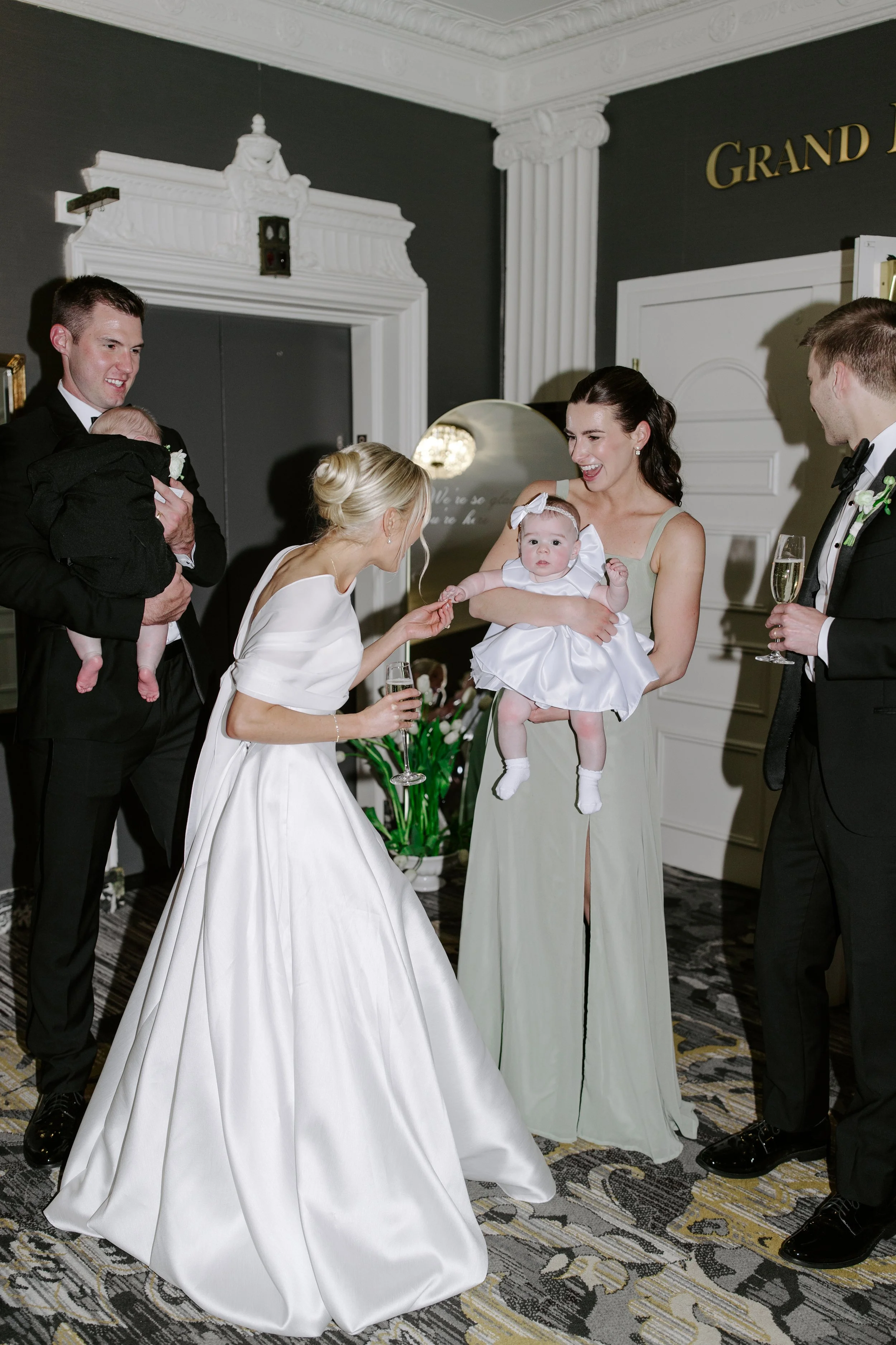 A wedding reception scene with a bride in a white gown, a woman handing her a glass, and a woman holding a baby, with men in tuxedos also present.