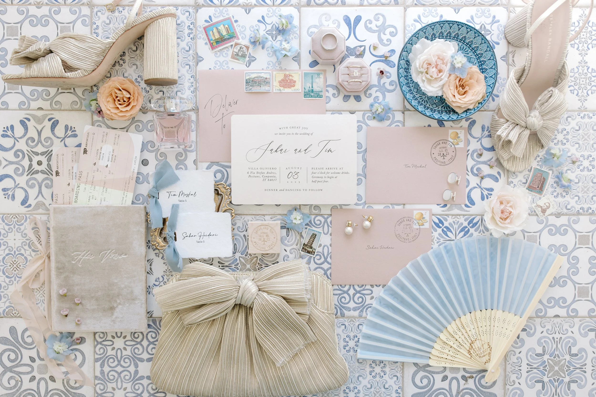 A collection of wedding invitation items and accessories arranged on a decorative tile surface. Includes wedding invitations, rings, tickets, jewelry, flowers, a fan, and decorative bows, with soft pastel colors.