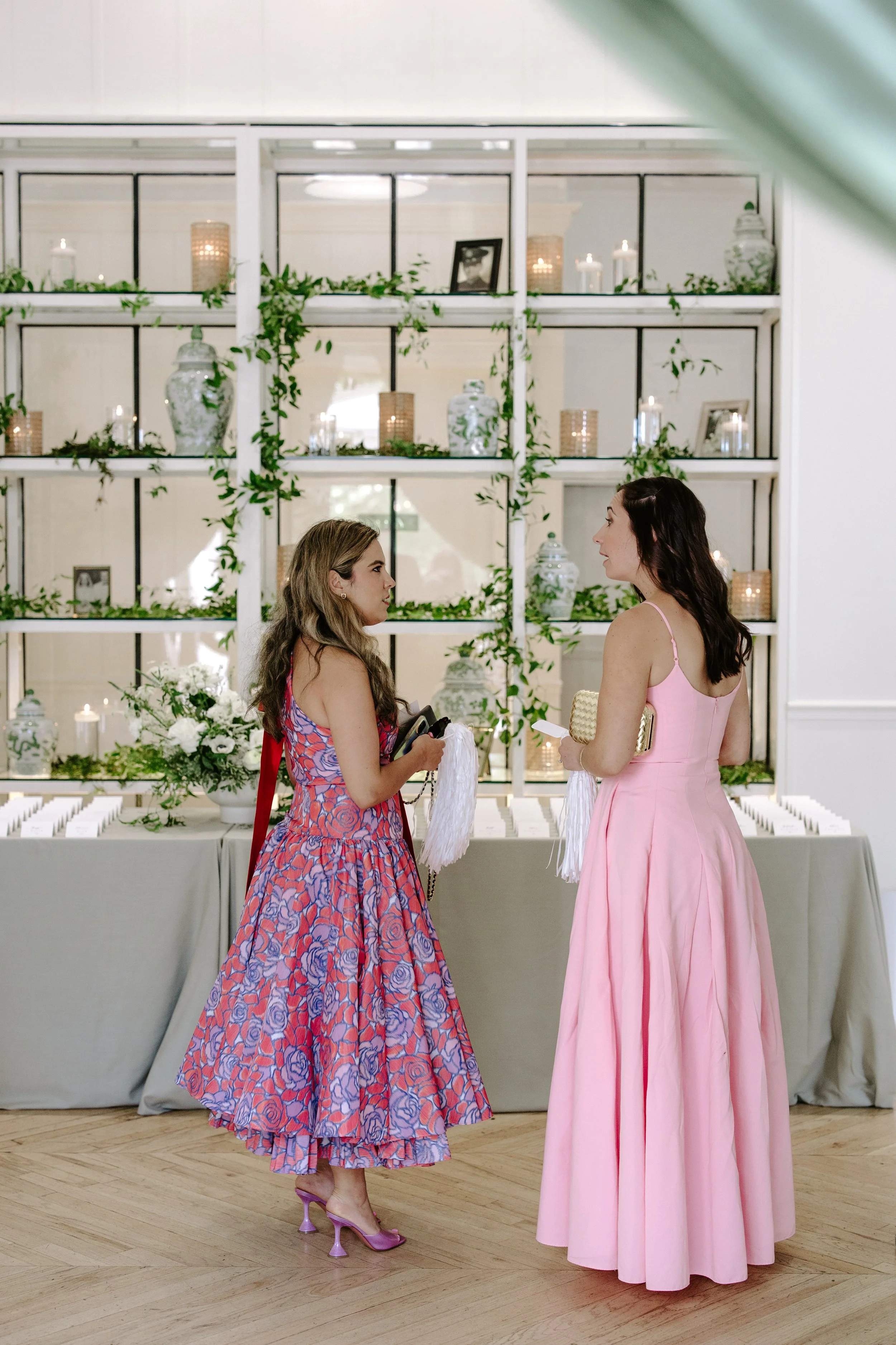 Two women in colorful dresses and heels face each other, exchanging items, in front of a decorated shelf with candles, vases, and framed photos, in a well-lit room with wood flooring.