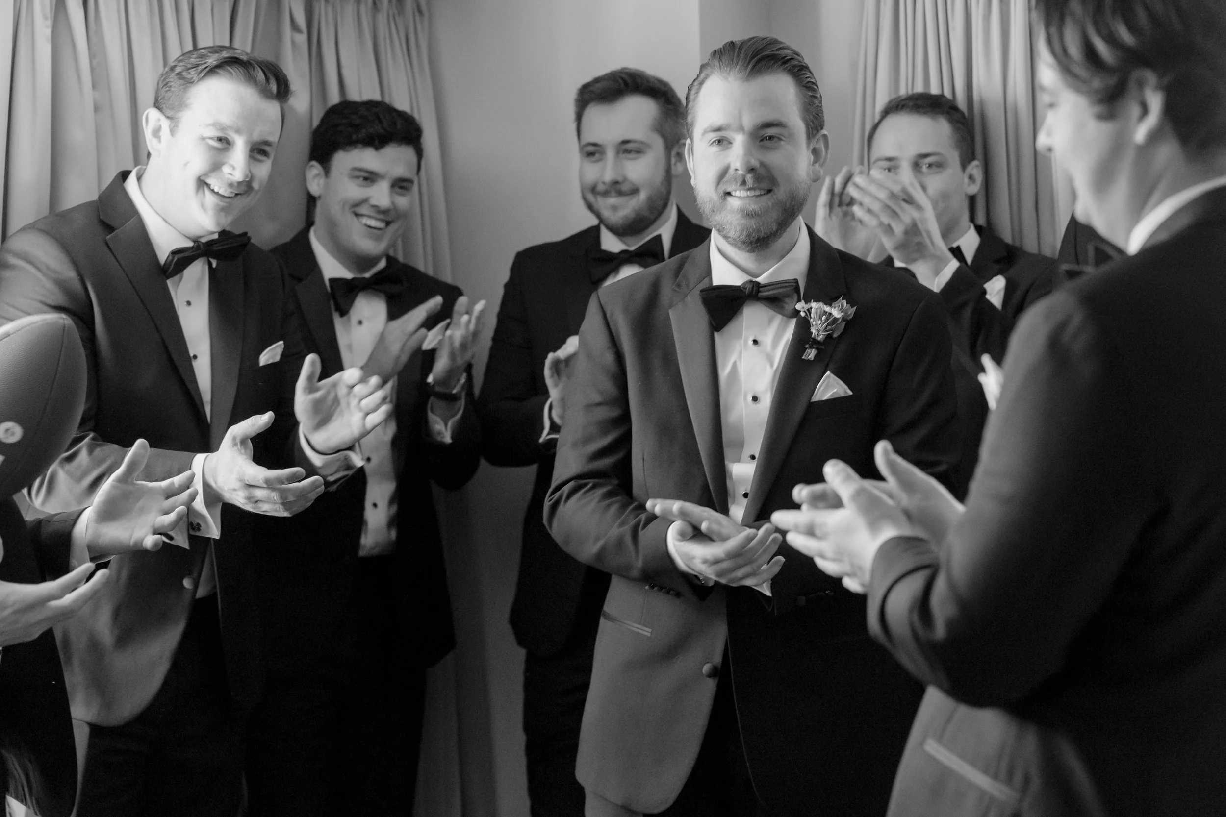 Six men dressed in tuxedos are gathered in a circle, smiling and clapping, in a celebratory setting, with curtains in the background.