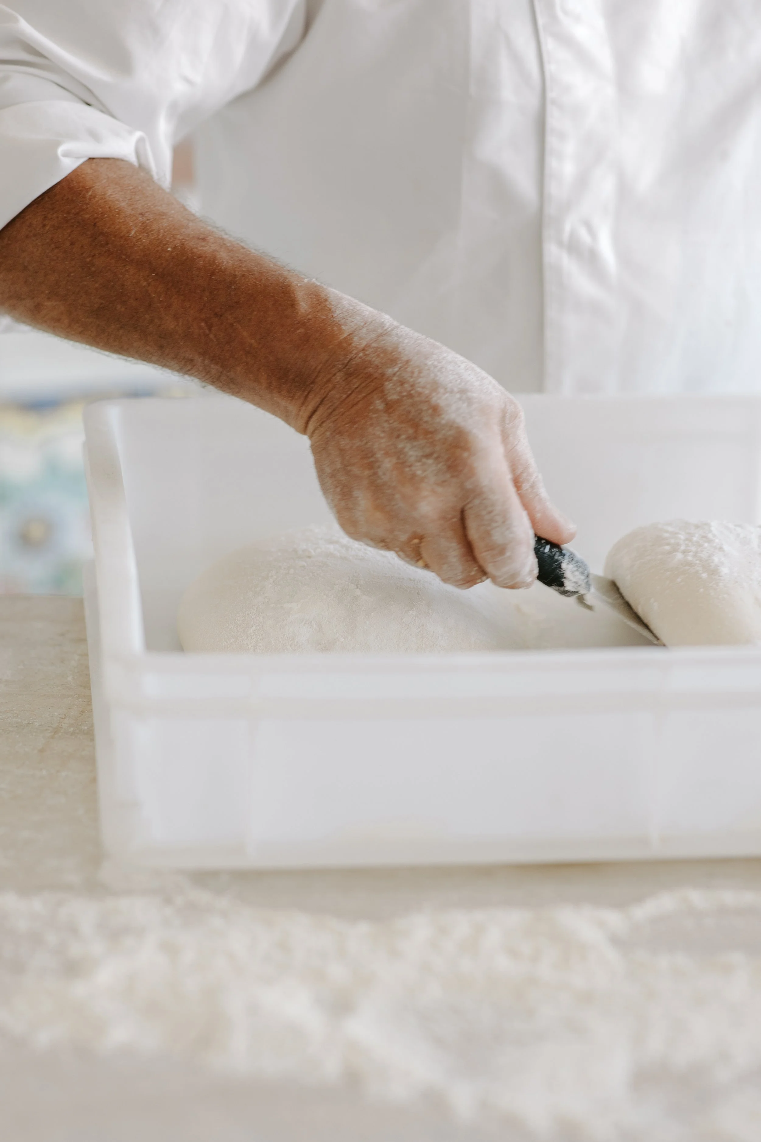 A person wearing a white shirt sprinkling flour onto a dough with a small black utensil in a white container.