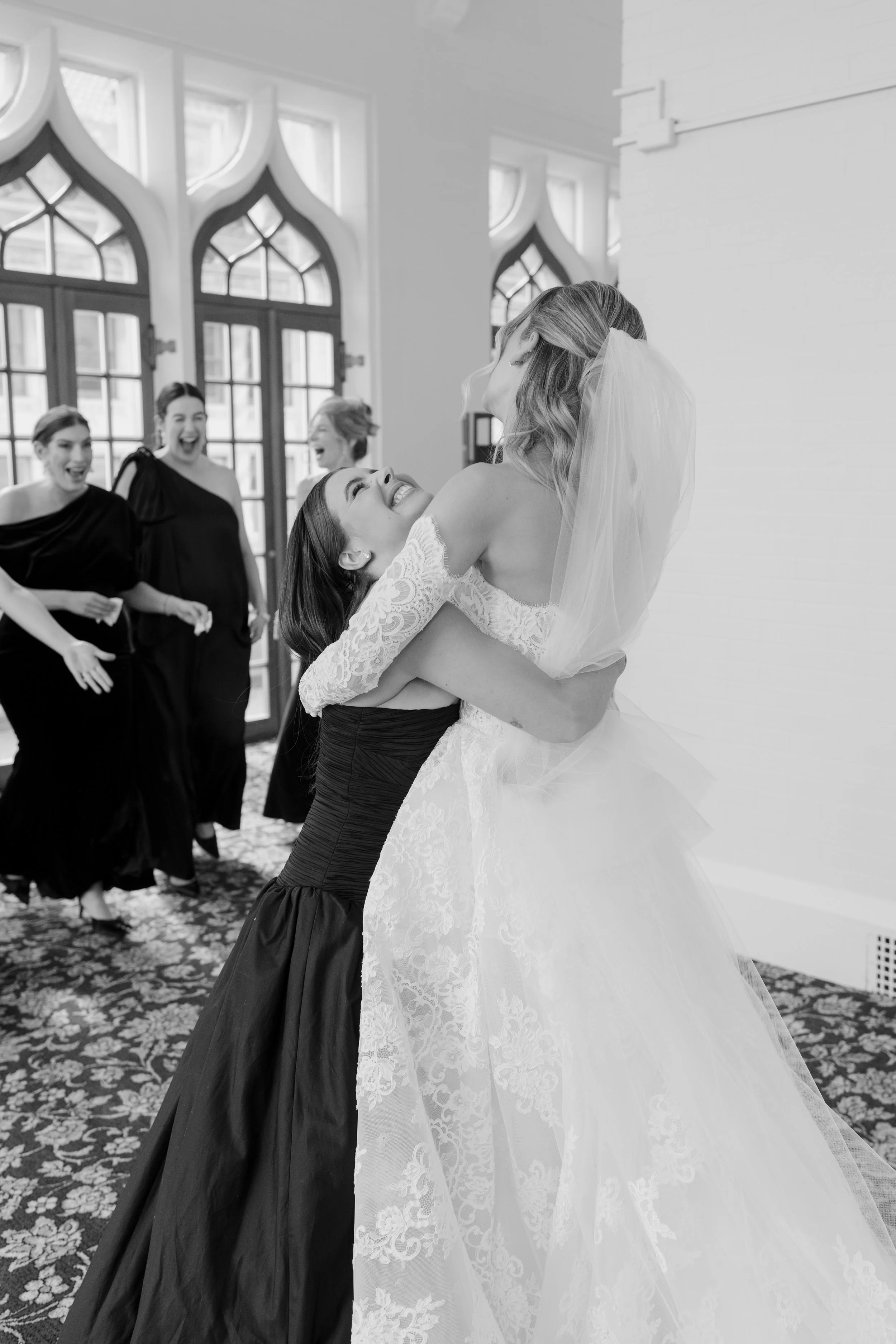 A bride in a wedding dress and veil being lifted up by a bridesmaid in a black dress, with three women in black dresses laughing in the background.