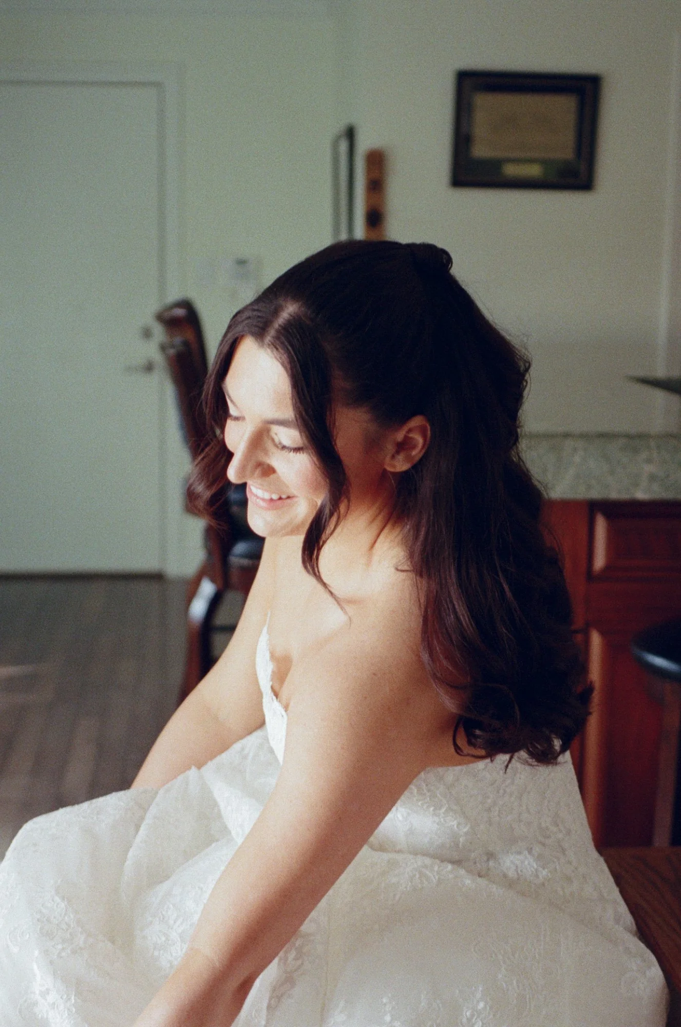 A woman with long, dark, wavy hair smiling while looking down, wearing a white lace dress, sitting in a room with a door and a framed picture on the wall.