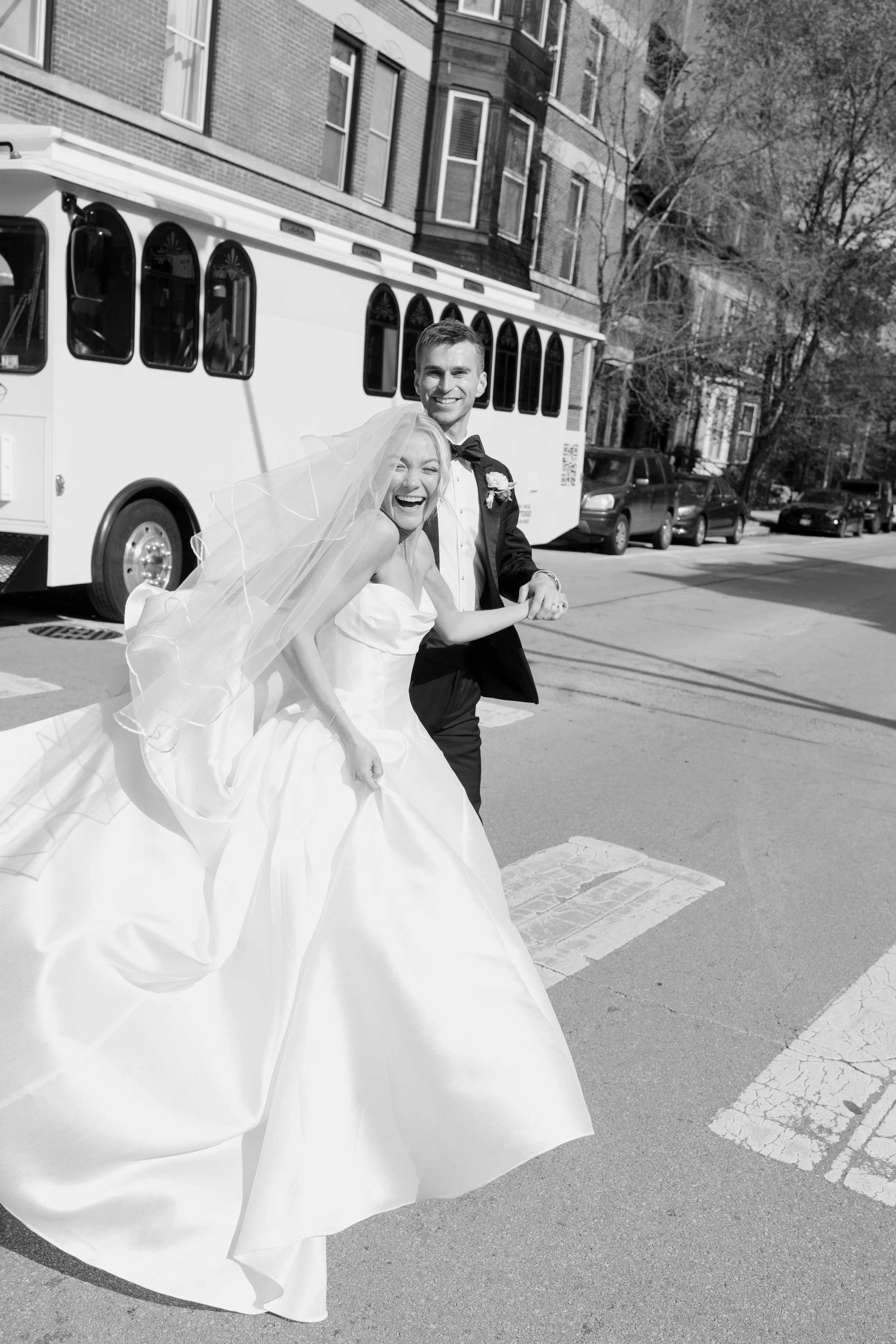 A smiling bride and groom holding hands and dancing in the street on a sunny day. The bride is wearing a wedding dress with a veil, and the groom is in a tuxedo with a bow tie. In the background, there is a streetcar, parked cars, trees, and brick bu