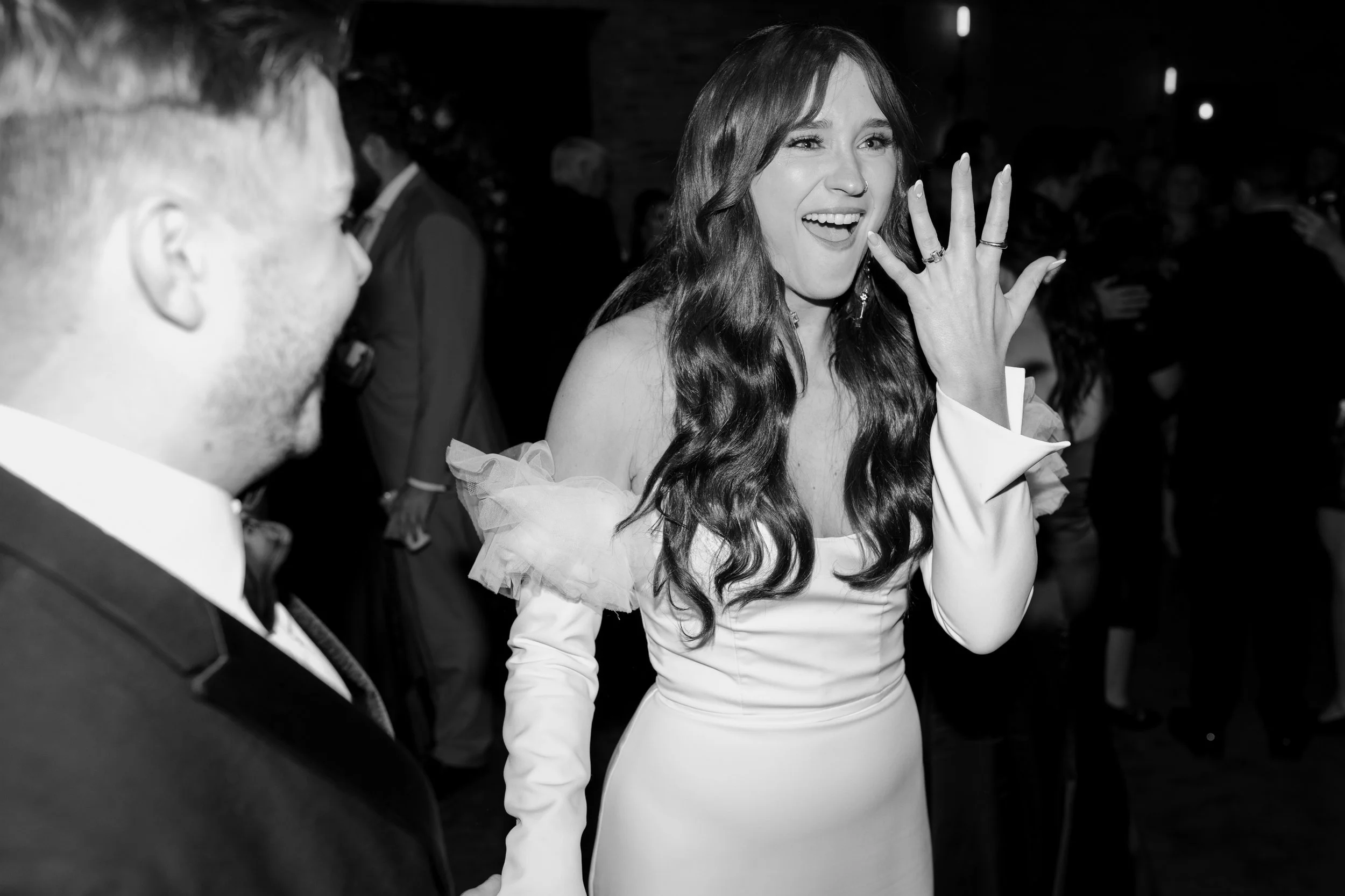 A woman with long wavy hair in a white dress showing her ring finger covered in rings, smiling and waving at a social event, with other people in the background.