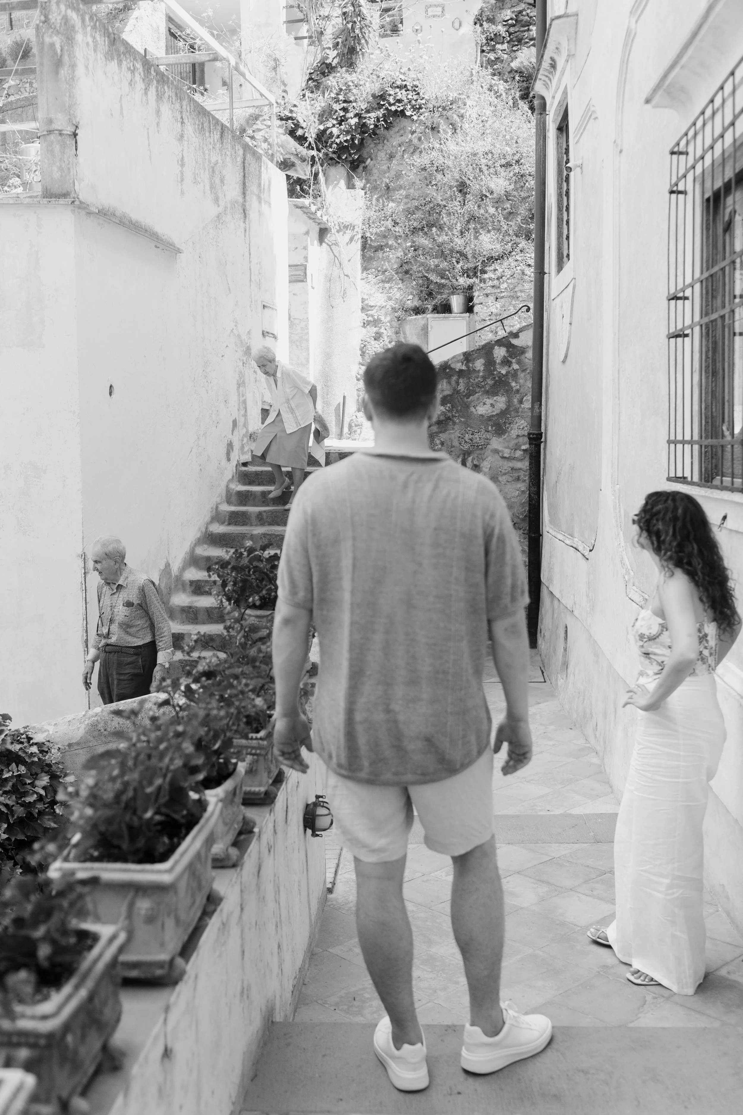 A man in a T-shirt and shorts standing on a street, observing an elderly woman walking up stairs. There are potted plants along a wall, and other people in the background, in an outdoor urban setting.