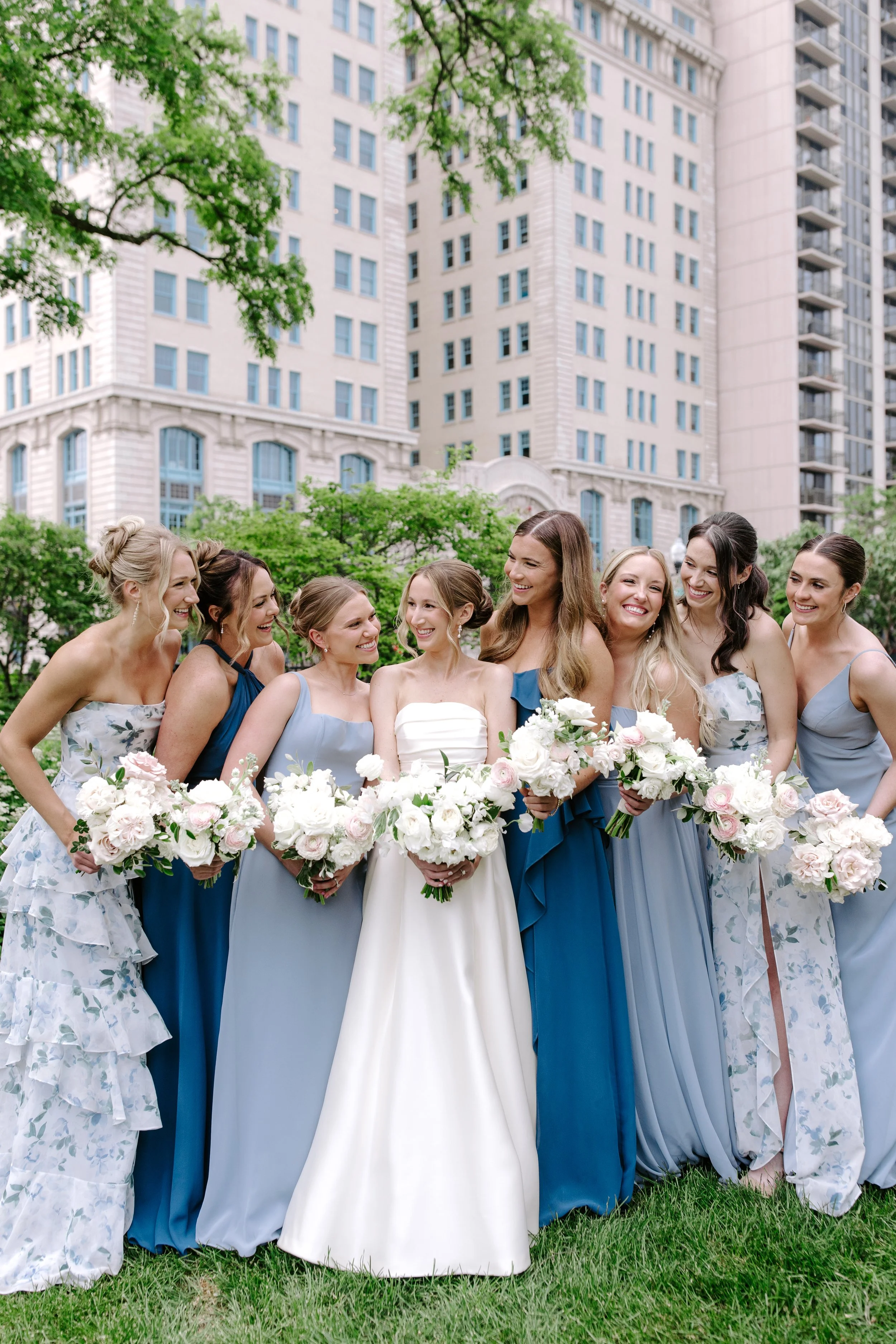 Group of women in dresses holding bouquets of flowers, standing together outdoors in front of tall buildings.