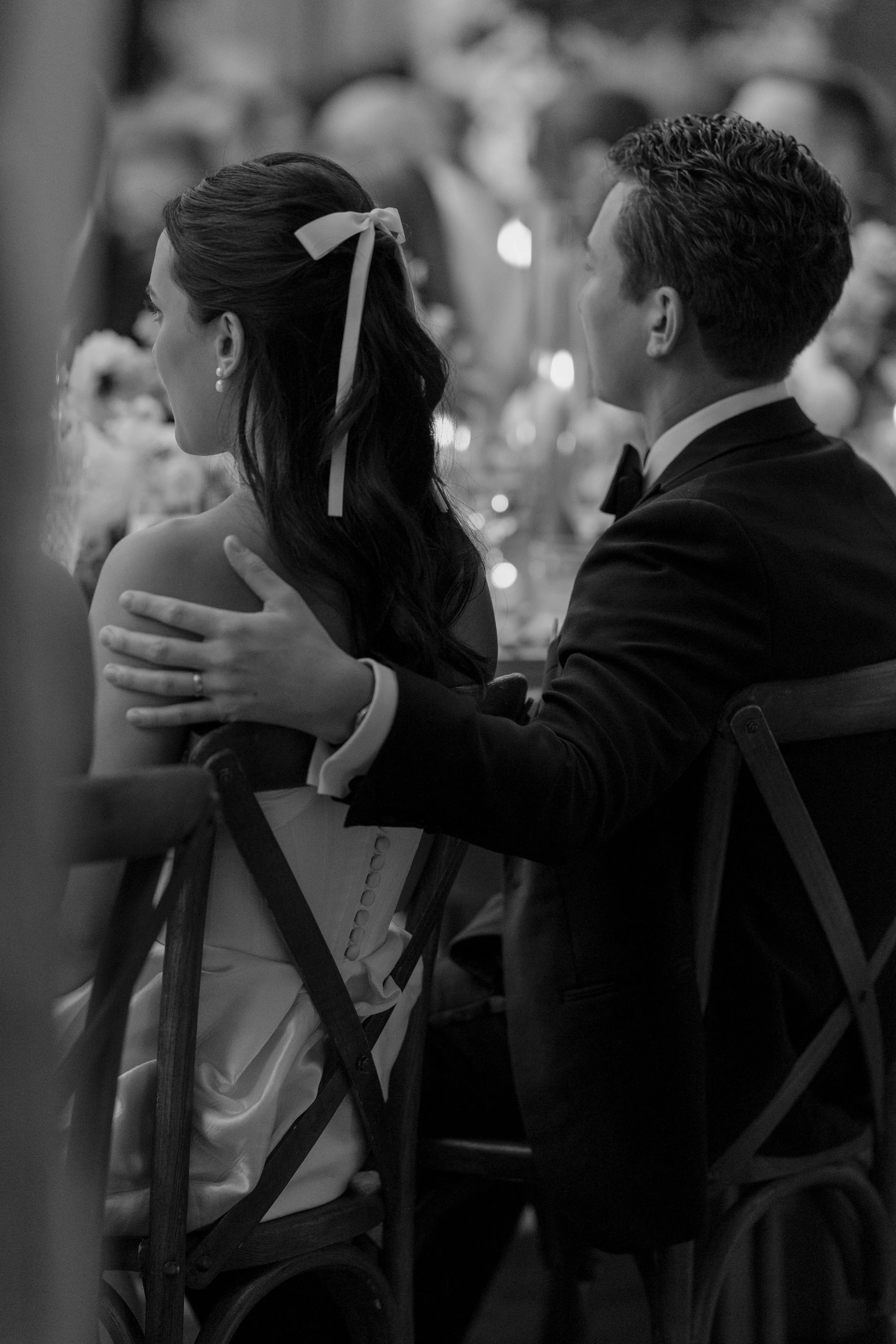 A couple dressed in formal attire sitting at a table, indicating a romantic or wedding setting, with blurred background of other guests.
