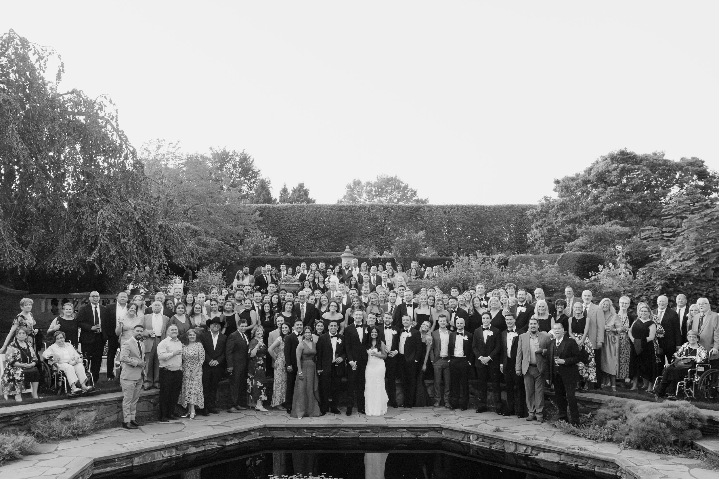 A large group of people in formal attire posing outdoors in a garden with trees and shrubbery, in front of a small pond.