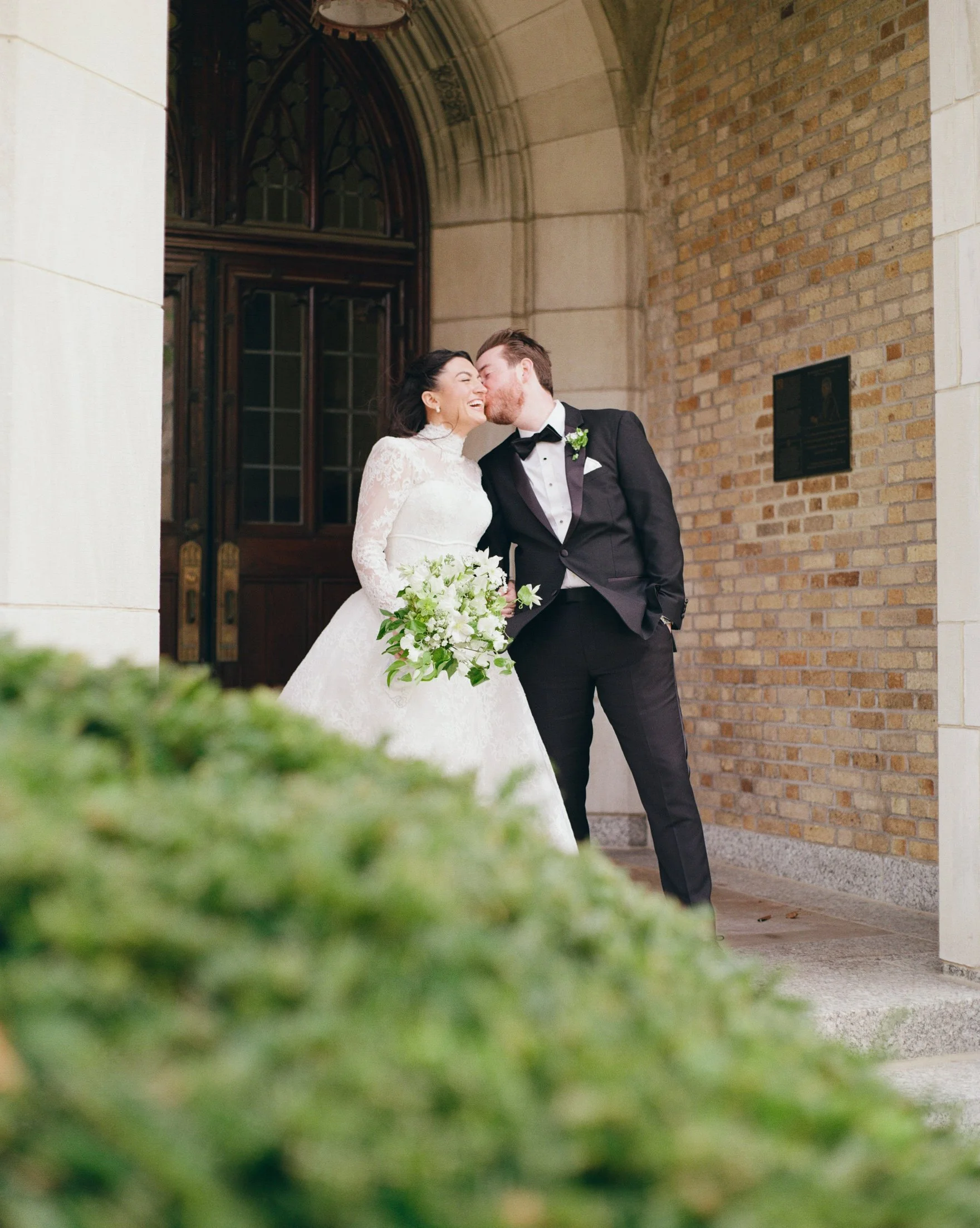 A bride and groom sharing a kiss outside a church or similar building, with the bride holding a bouquet of white flowers and wearing a white lace wedding dress, while the groom is dressed in a black tuxedo with a bow tie and boutonniere, against a ba