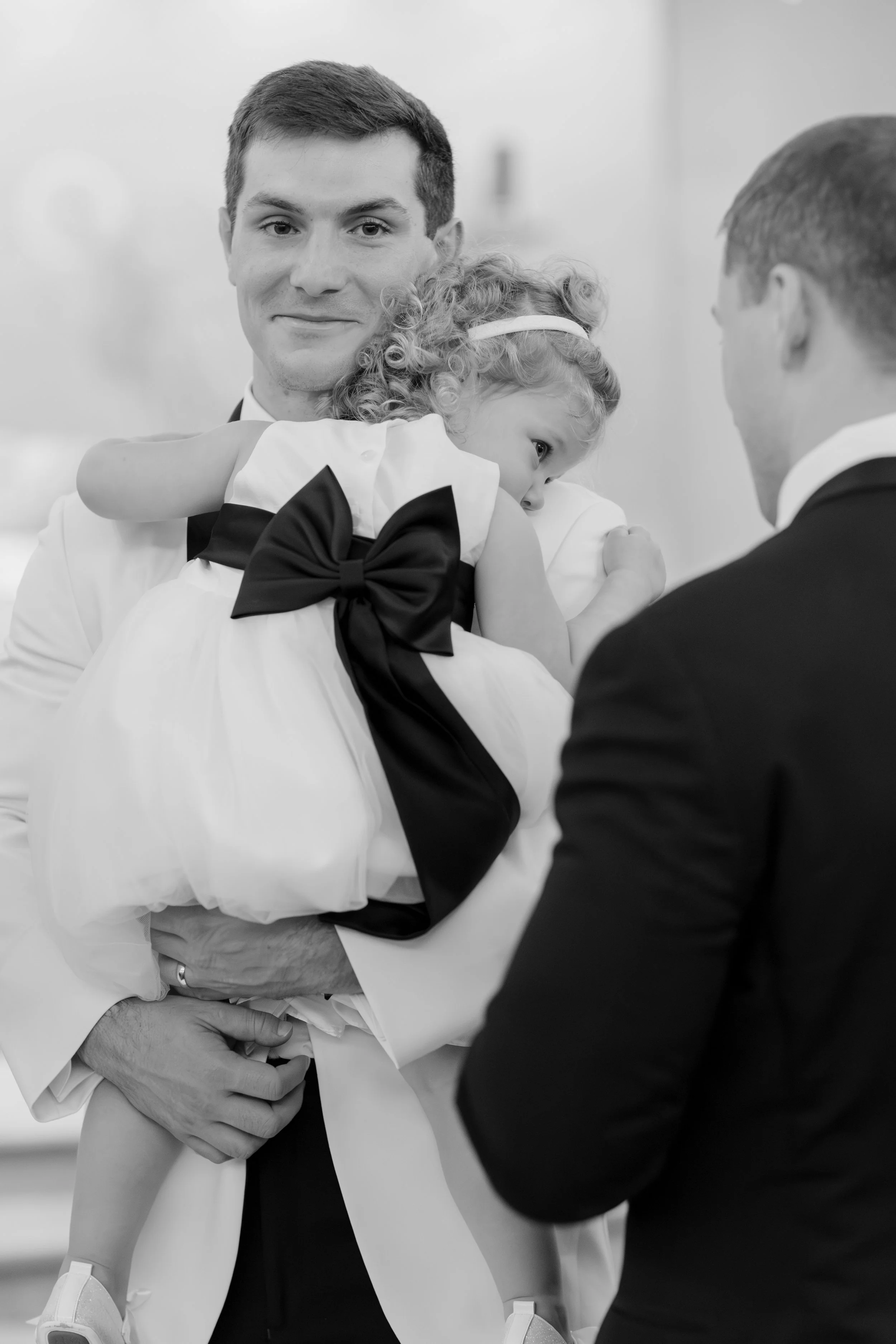 A man in a suit holding a young girl with curly hair and a headband, embracing her while a second man in a suit looks on. The photo is in black and white.