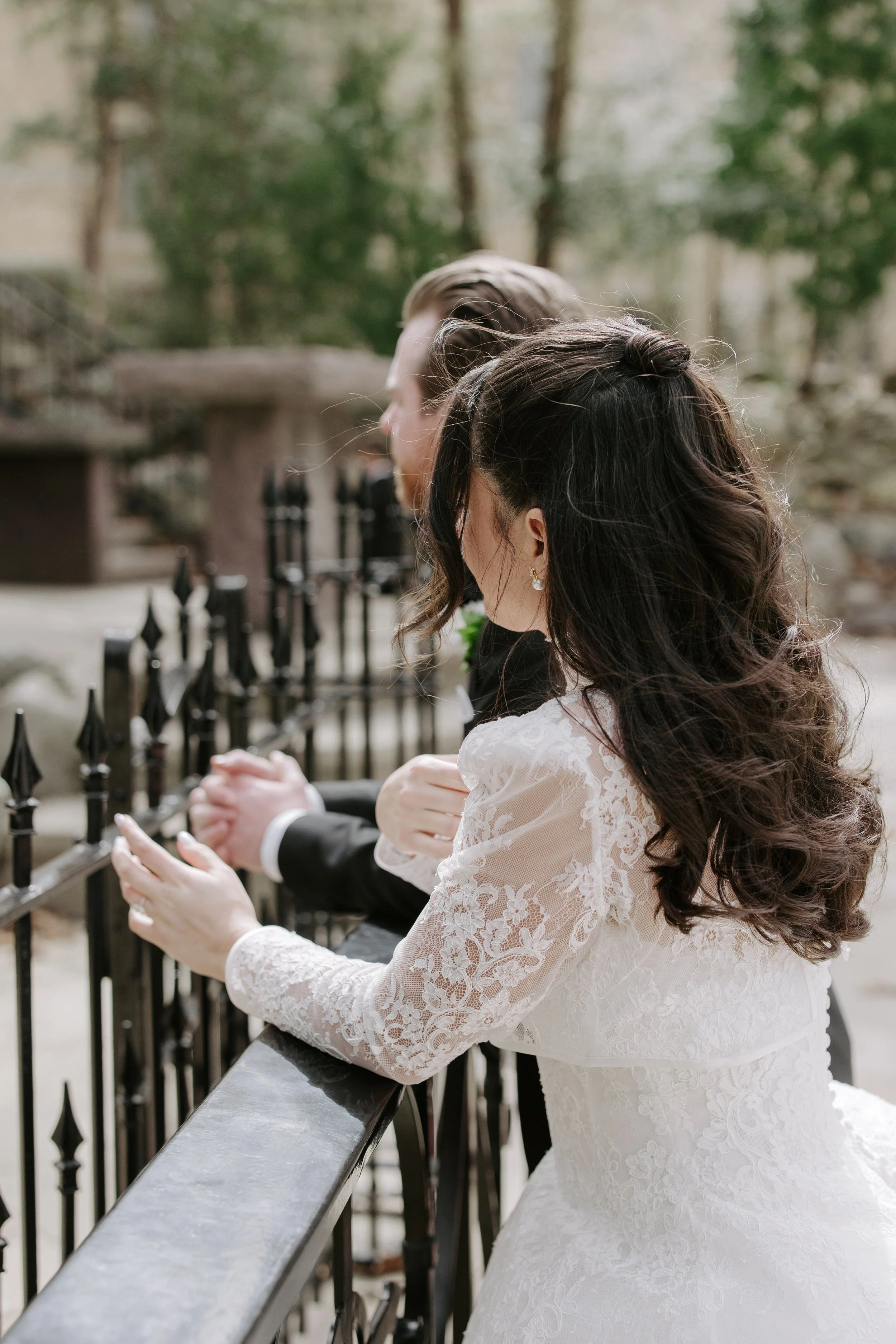 A bride and groom are praying together outdoors, holding hands on a stone railing with a black metal fence behind them, surrounded by trees.