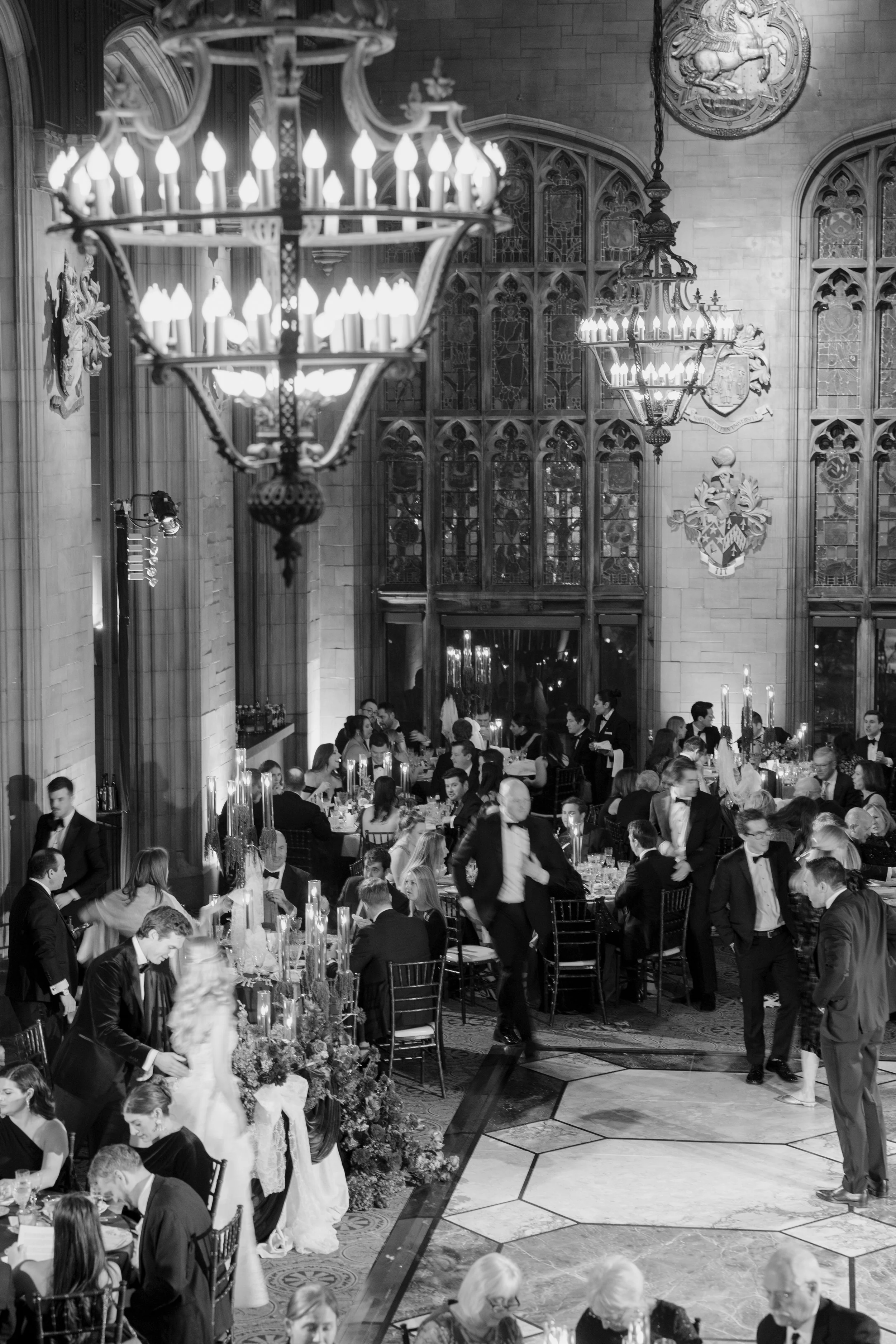 A formal dinner event in a grand hall with chandeliers, stained glass windows, and guests in tuxedos and evening gowns seated at decorated tables.