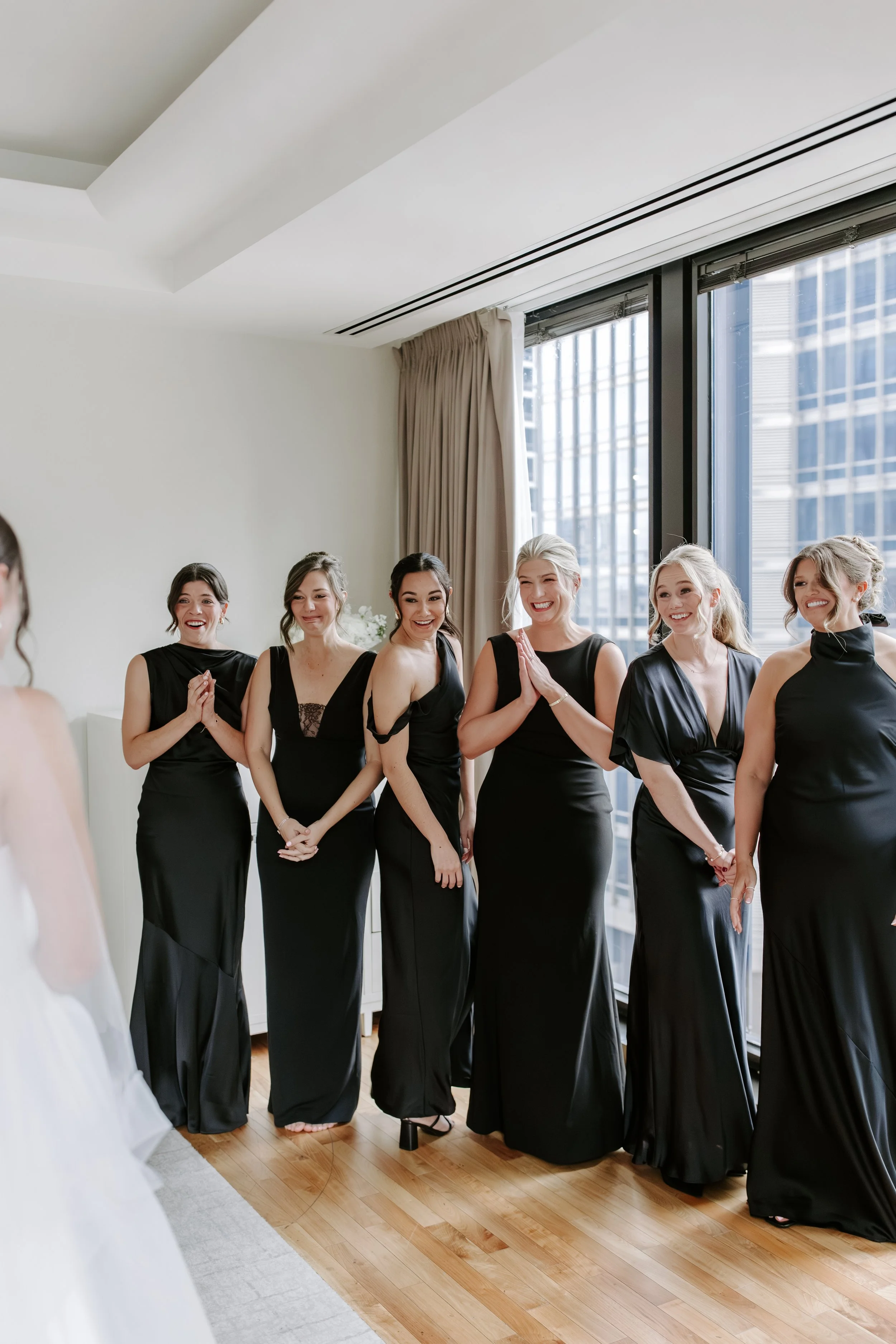 A group of women in black dresses standing in a line indoors near large windows, smiling and enjoying a moment together.