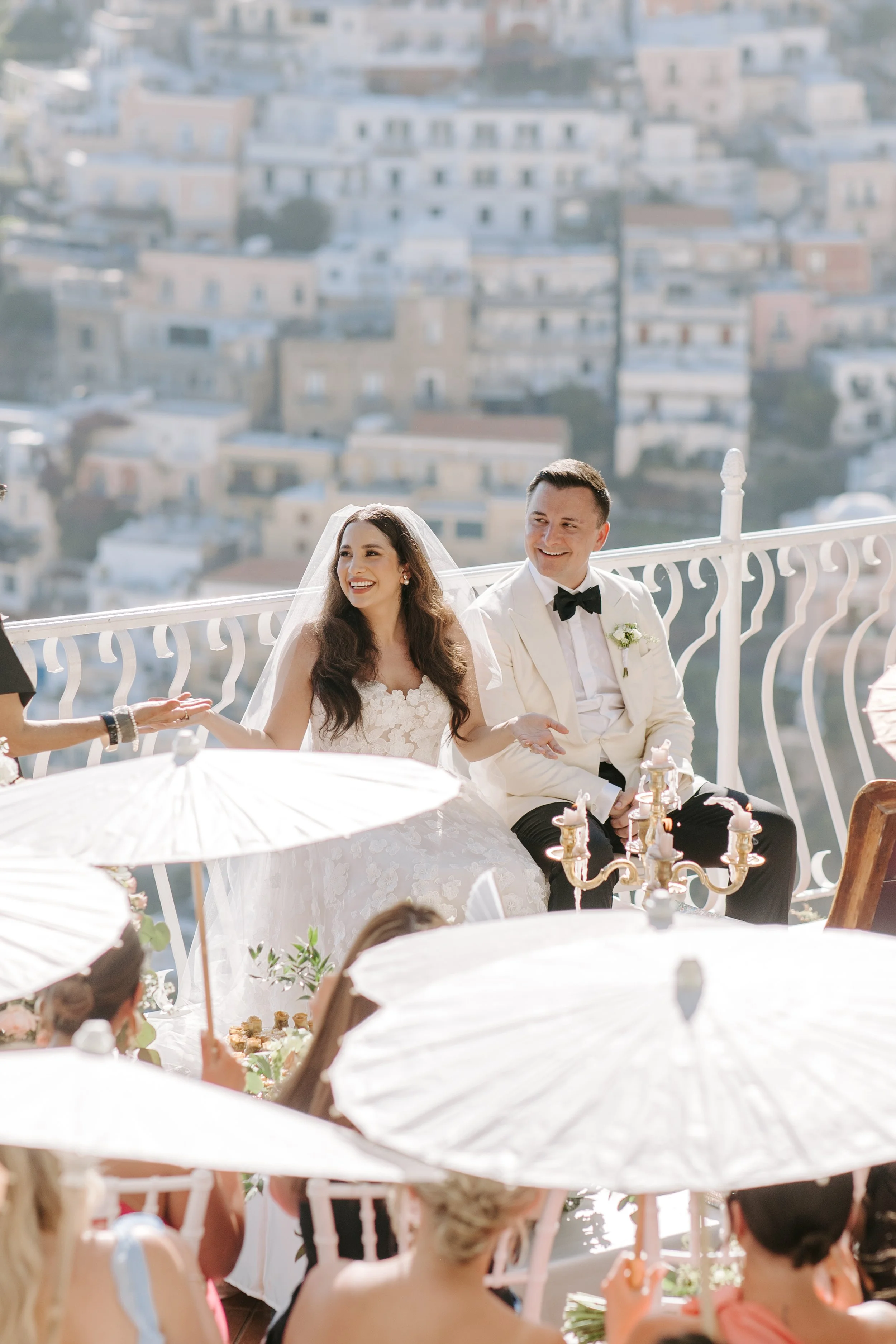 A bride and groom sitting at a wedding reception, smiling amidst guests holding white parasols, with a view of a hillside cityscape in the background.