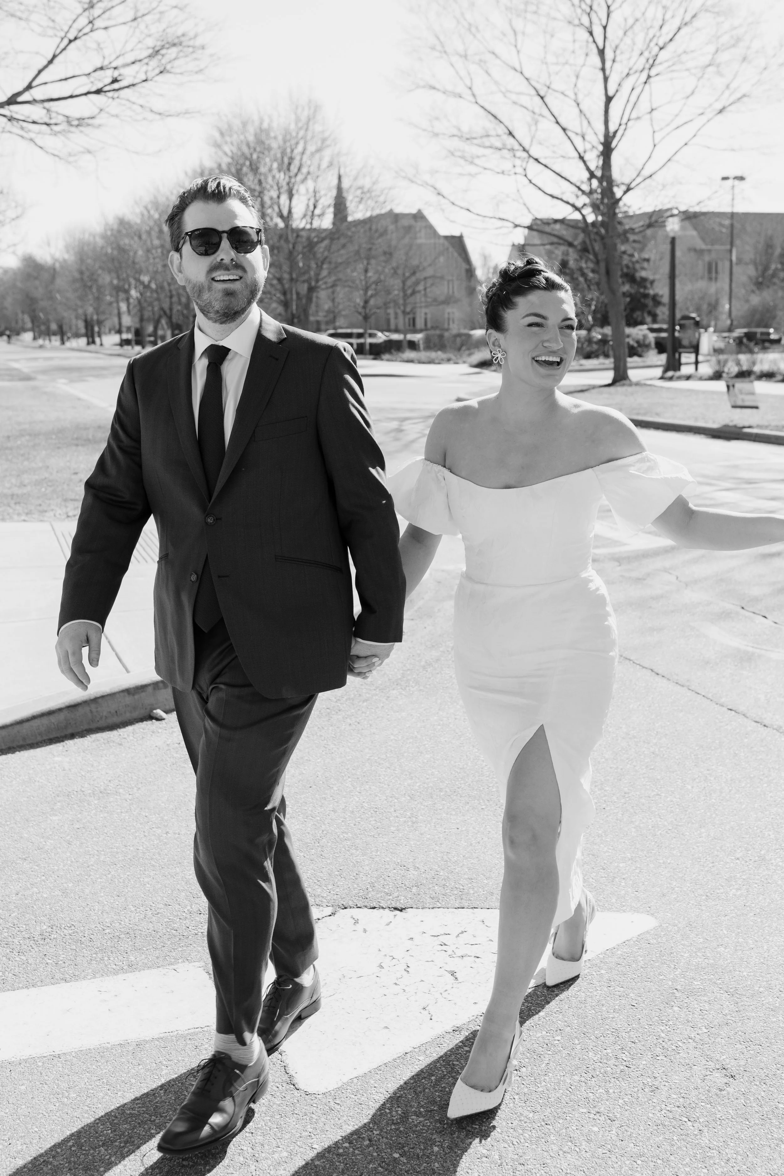 A black-and-white photo of a smiling man and woman walking hand in hand outside on a sunny day, with leafless trees and buildings in the background.