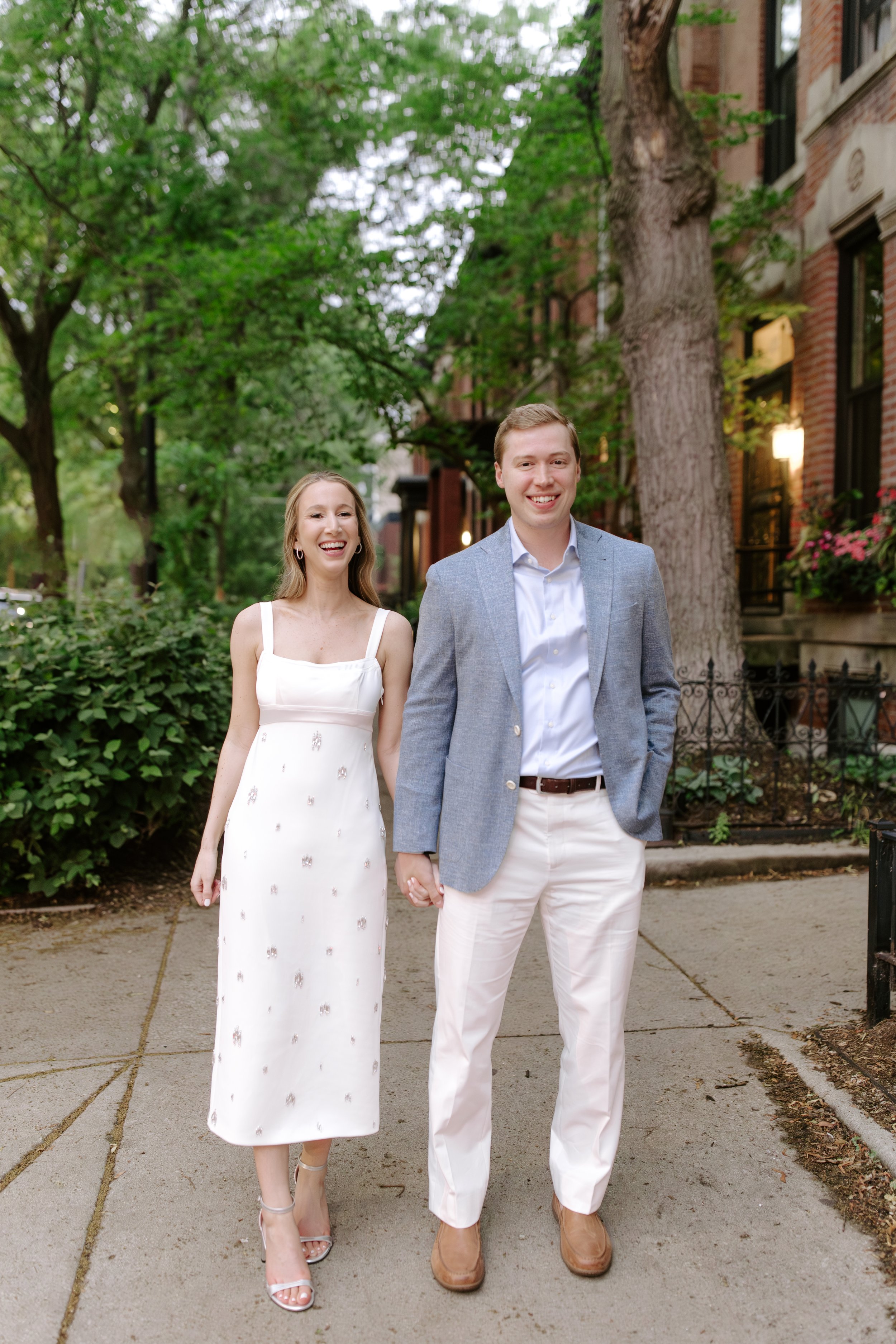 A smiling couple holding hands and walking on a sidewalk outdoors, surrounded by green trees and a brick building in the background, dressed in semi-formal attire.