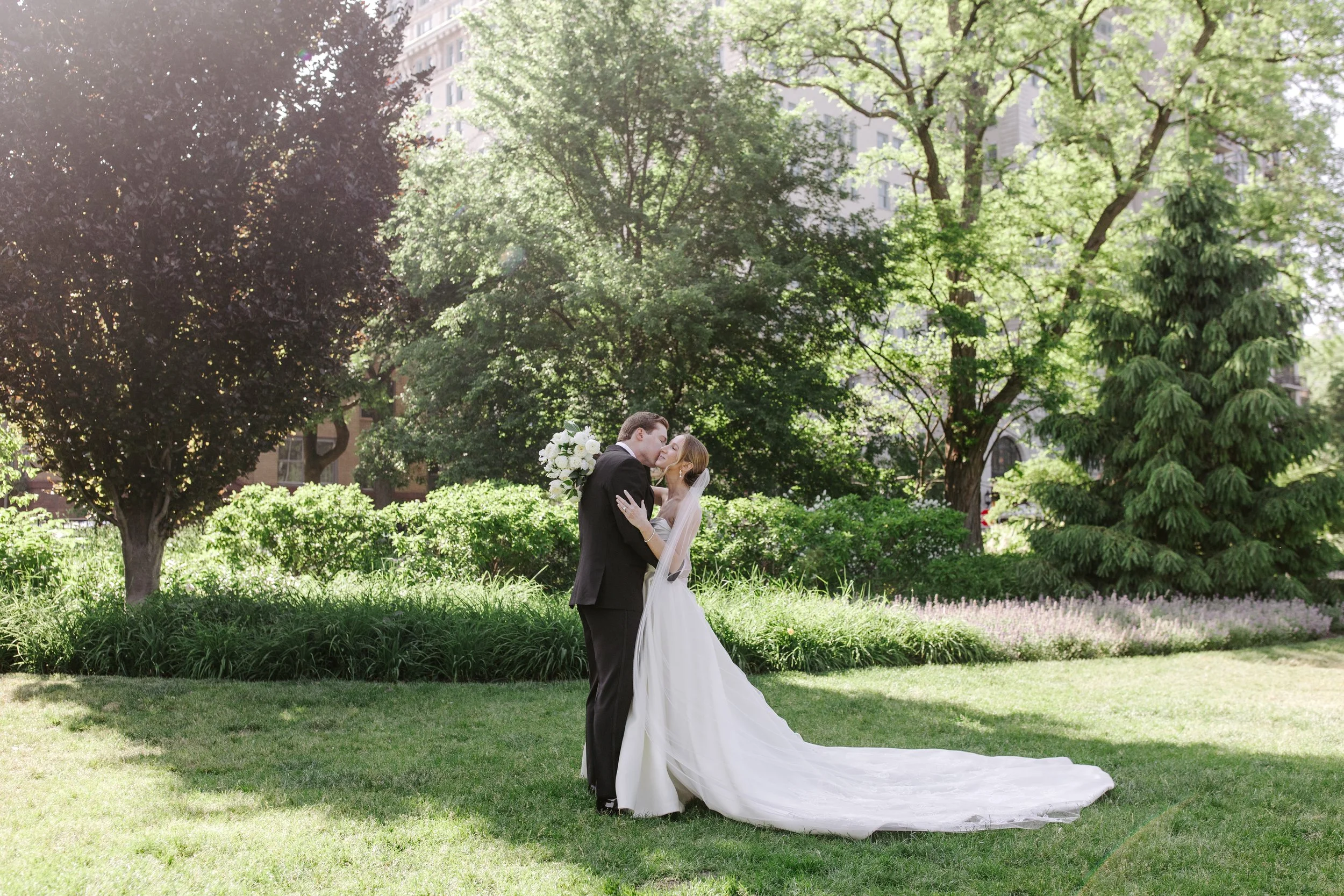 A bride and groom sharing a kiss in a park with green trees and buildings in the background.