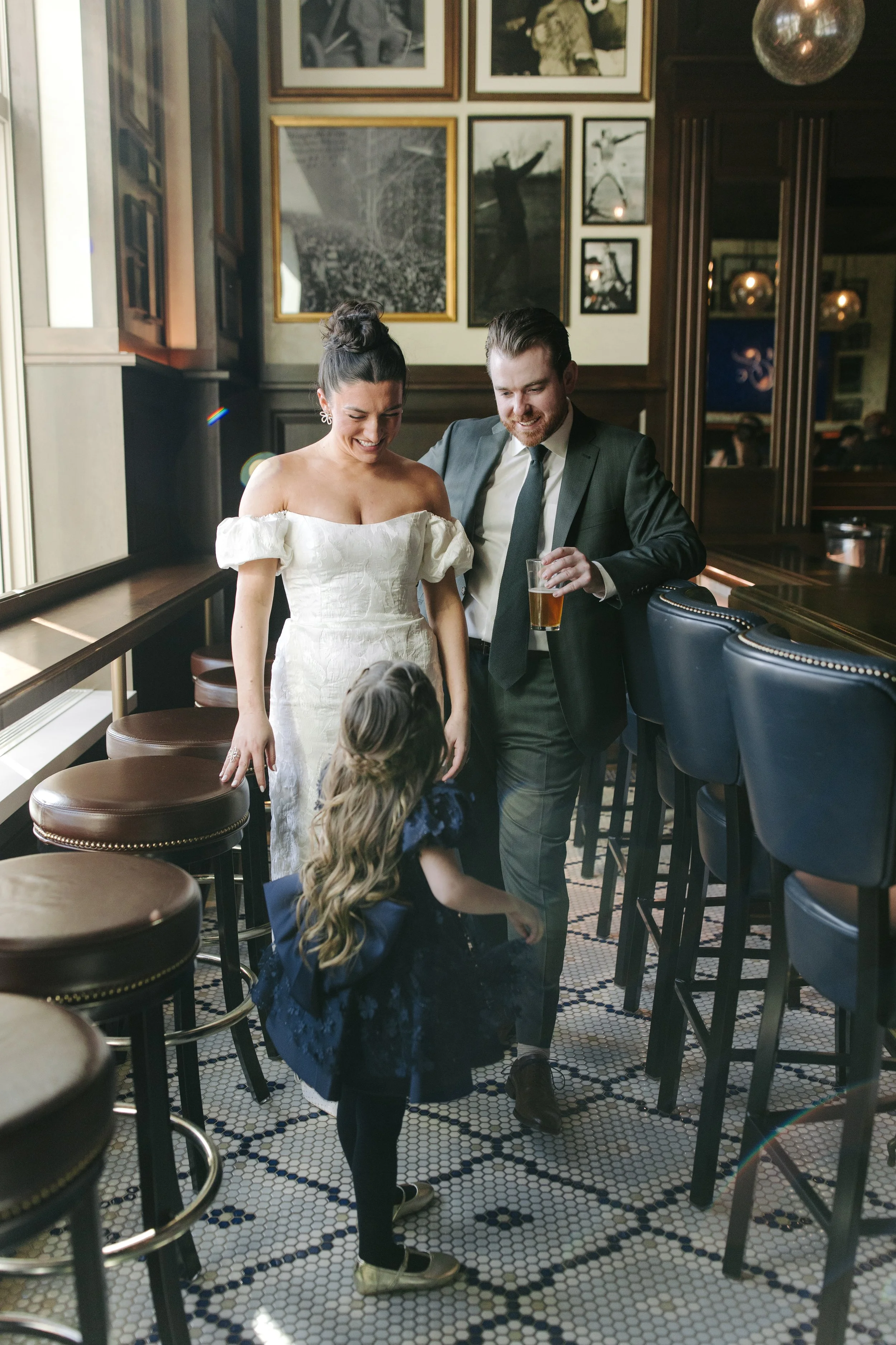 A woman in a white off-shoulder dress and a man in a suit are smiling and talking to two young girls with long hair inside a restaurant with dark wood paneling, black and white photographs on the wall, bar seating, and patterned tile floor.