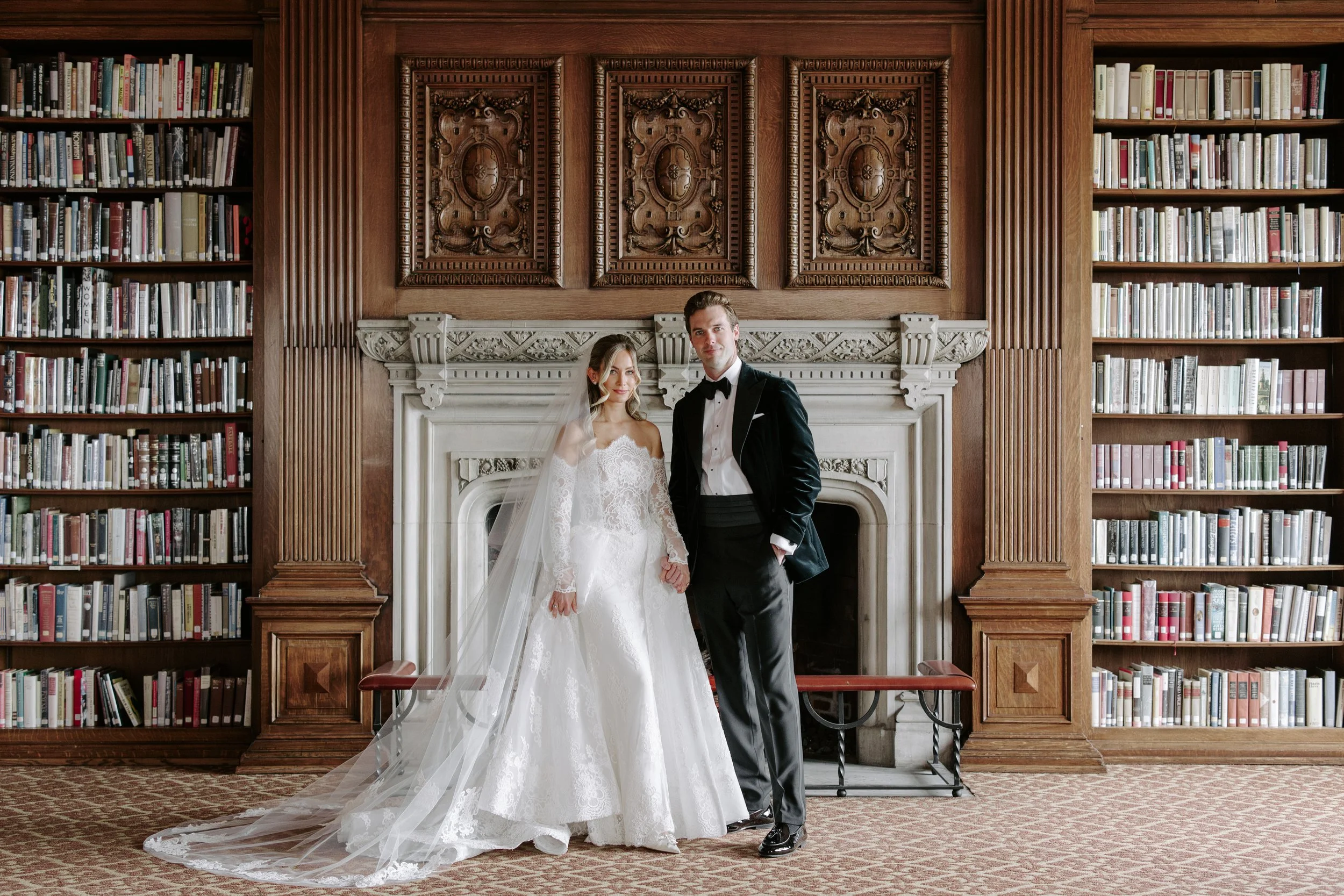 A bride and groom pose in front of a ornate wooden fireplace with carved details, inside a library with tall bookshelves filled with books.