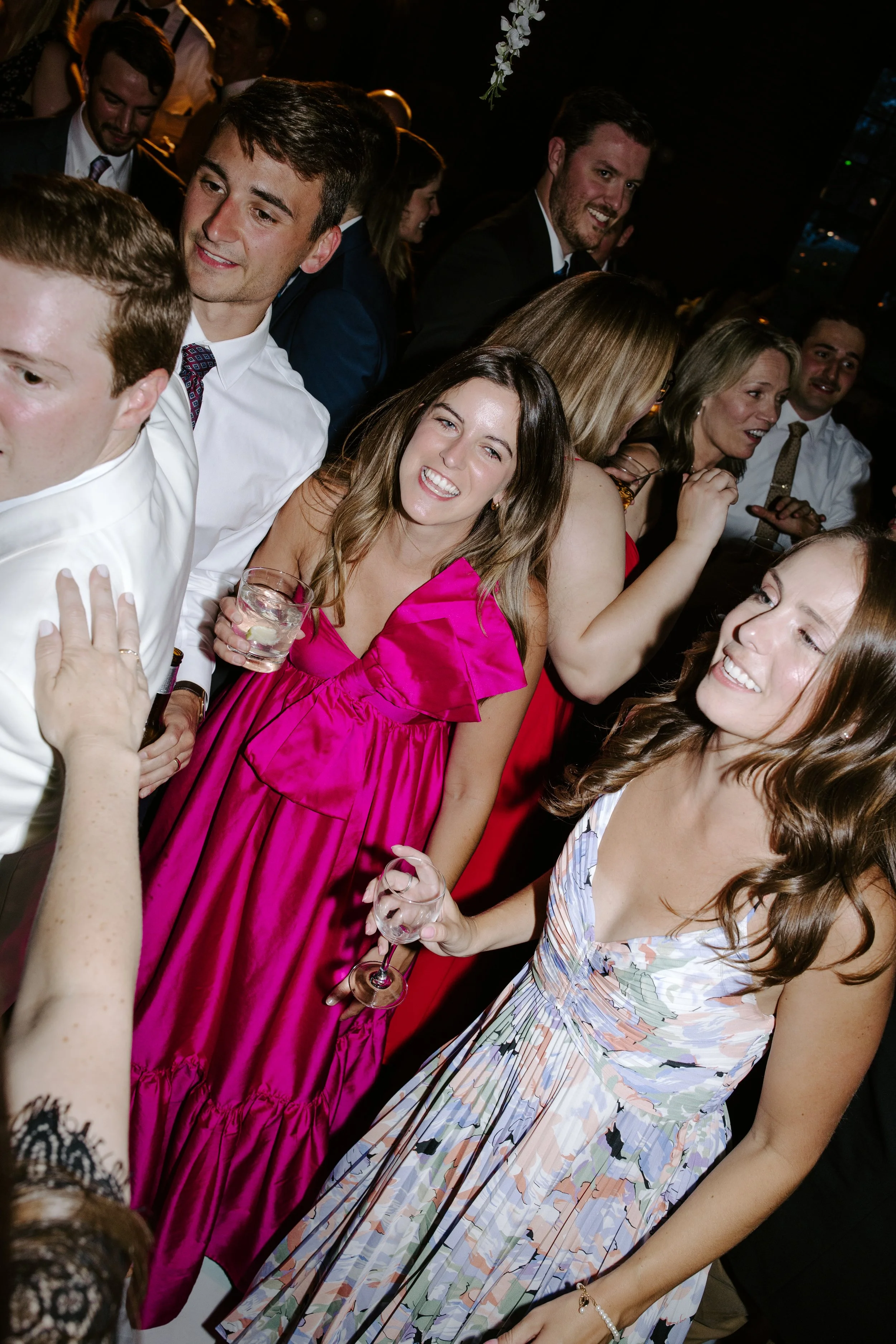 People socializing at an indoor event, some holding glasses of wine, wearing formal and semi-formal attire, with smiles and engaged in conversations.