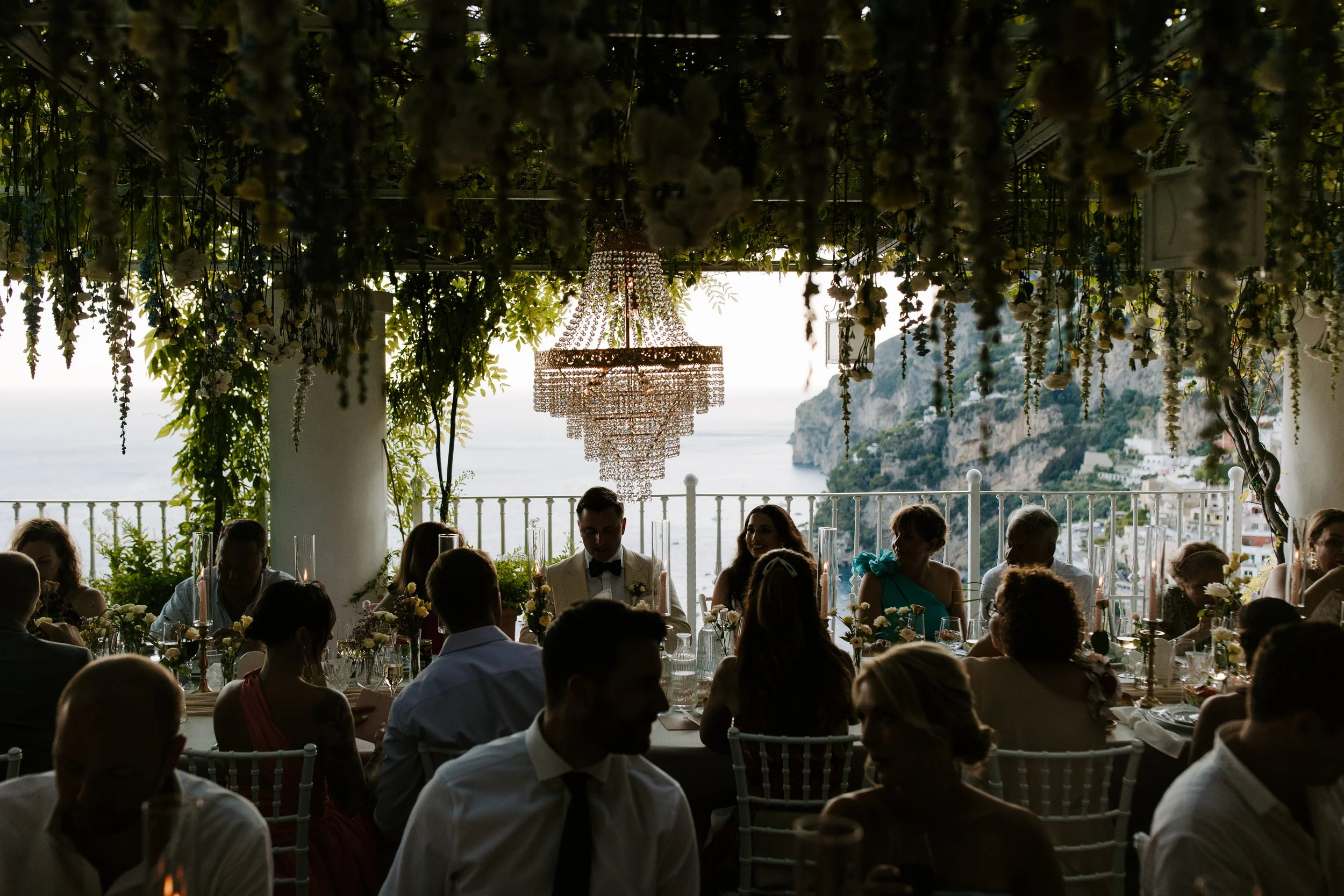 Indoor wedding reception with guests seated around tables decorated with flowers, overlooking an outdoor scenic view of cliff sides and water, with a chandelier hanging from a ceiling filled with hanging flowers and greenery.