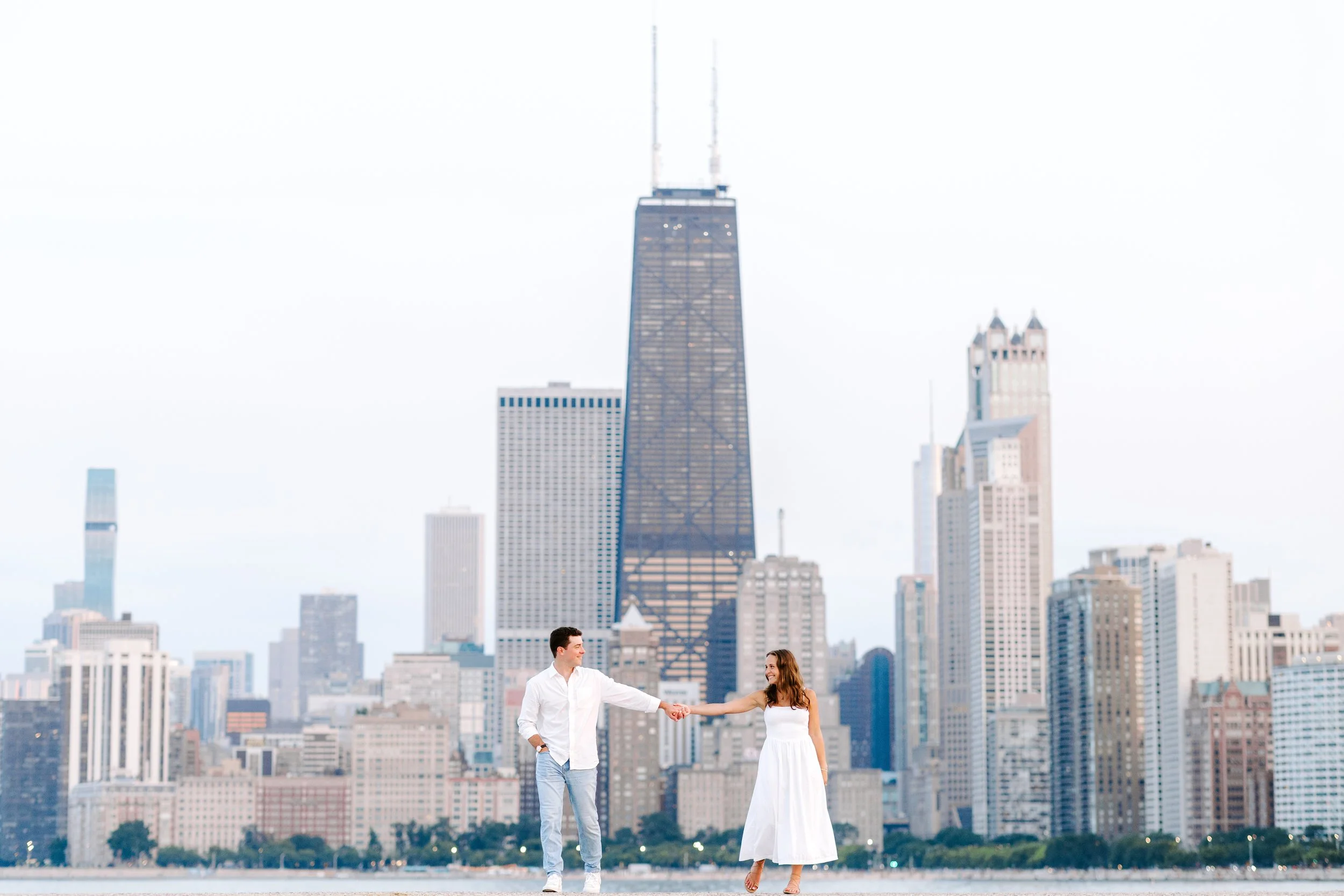 A man and woman holding hands and smiling with a city skyline in the background, including the tall building One World Trade Center, during daytime.