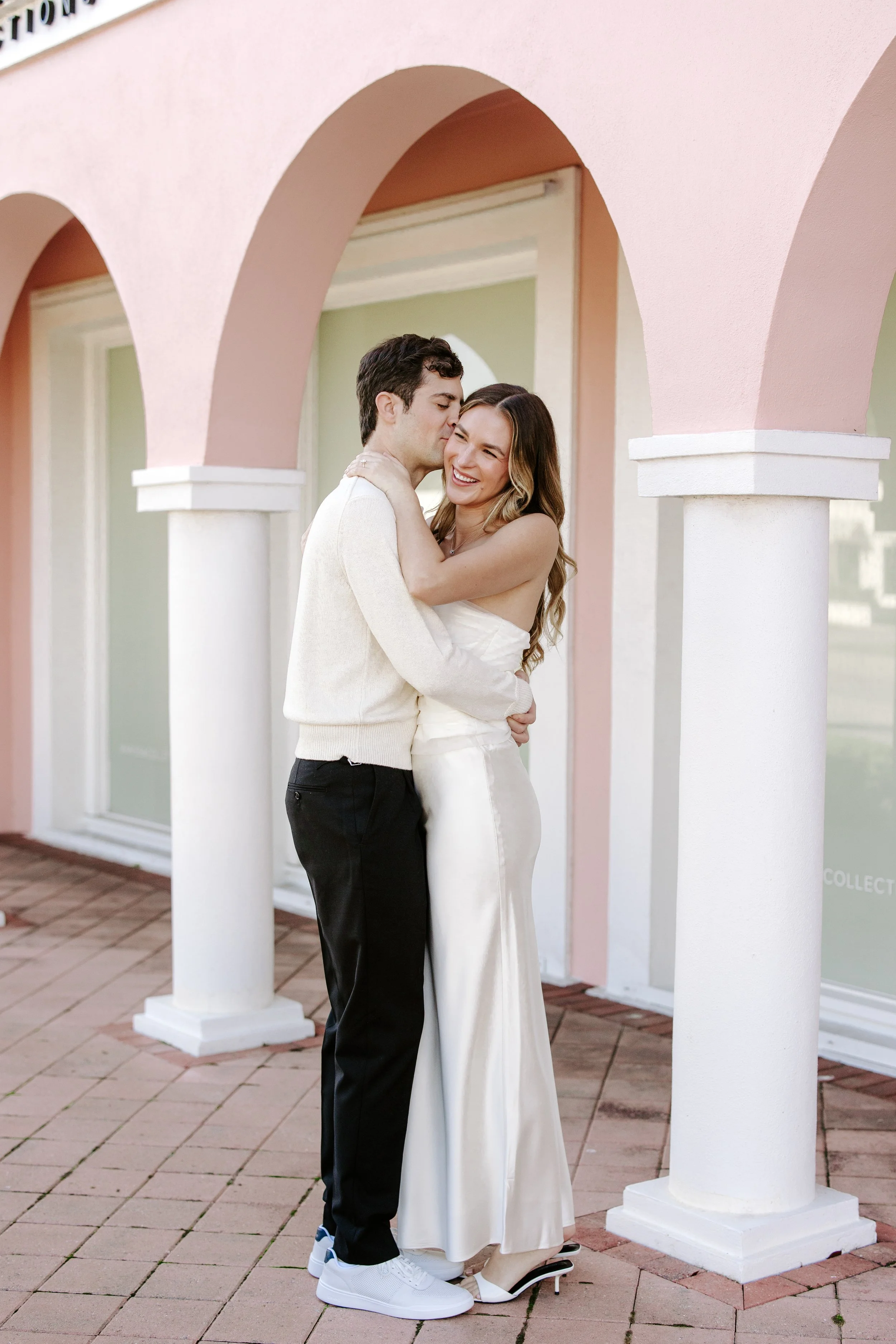 A couple hugging and smiling, standing under pink and green arches outside a building with large windows.