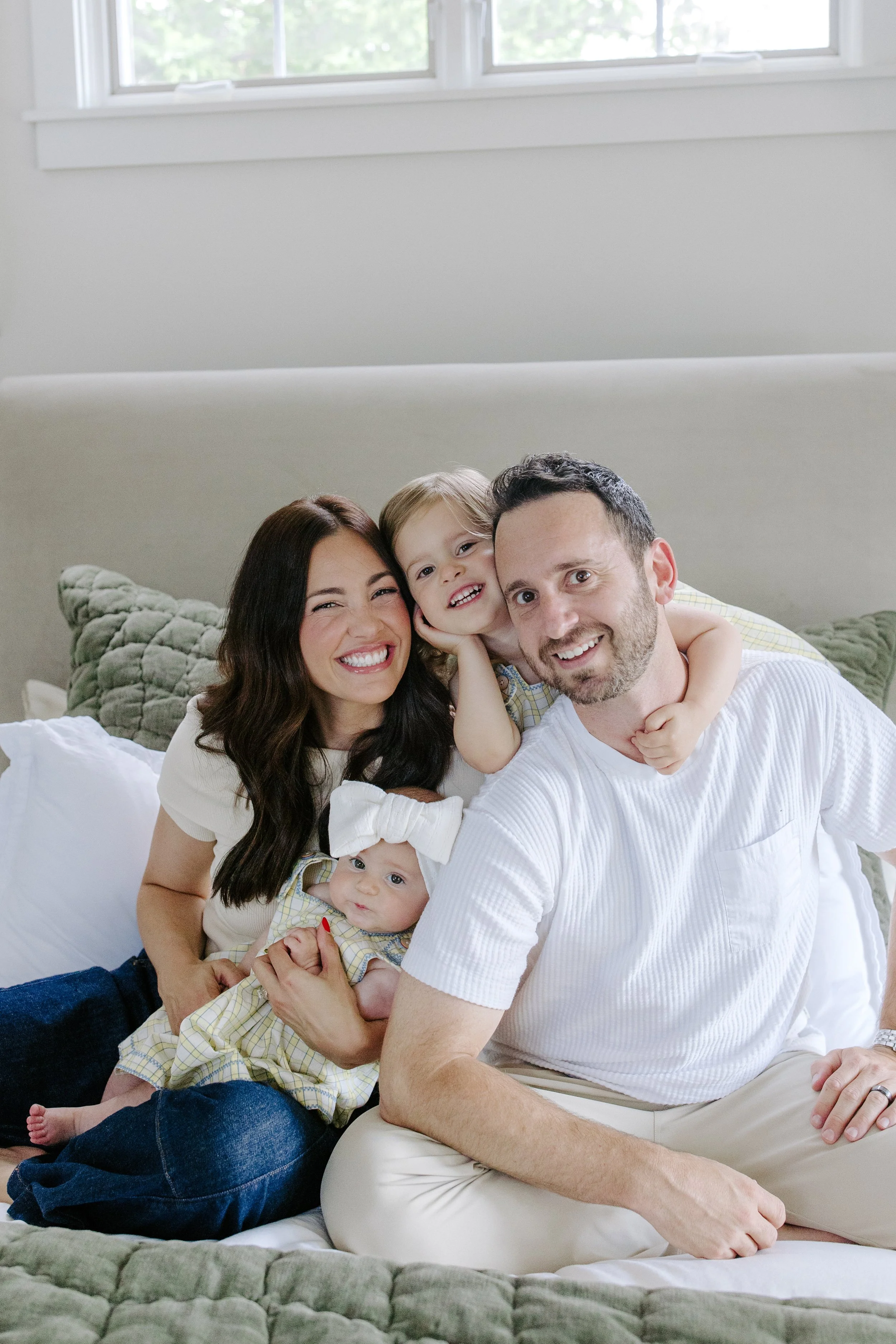A happy family of four, consisting of a woman, a man, and two children, sitting on a bed in a bright bedroom with a white wall and a window in the background.