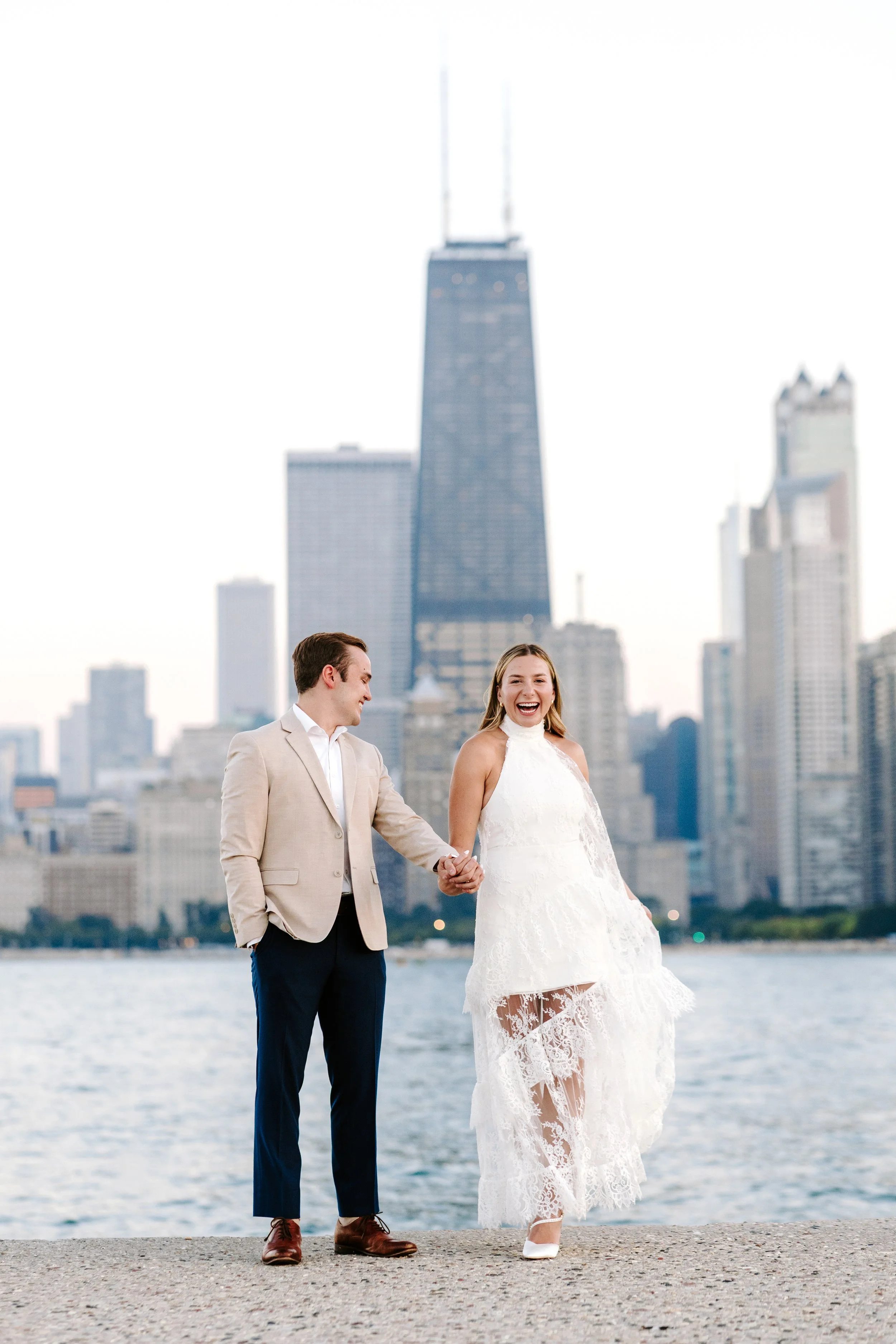 A bride and groom holding hands and walking along a waterfront with a city skyline, including the Willis Tower, behind them. The bride is smiling and wearing a lace wedding dress, while the groom is dressed in a beige suit with dark pants.