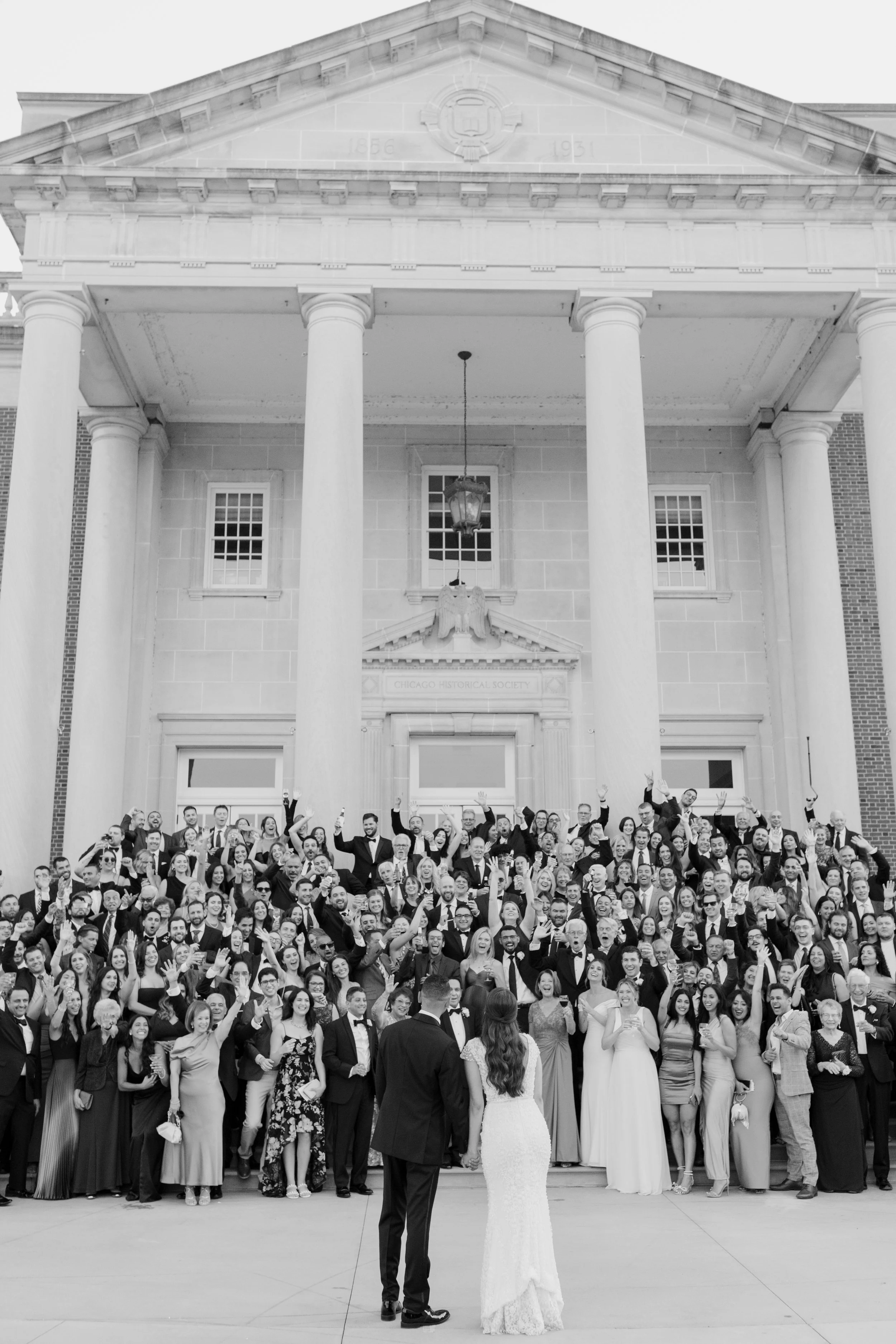 Black and white photo of a large wedding party posing on the steps of a historic building with grand columns and a triangular pediment. The bride and groom are in the foreground, facing away from the camera, with the crowd cheering behind them.