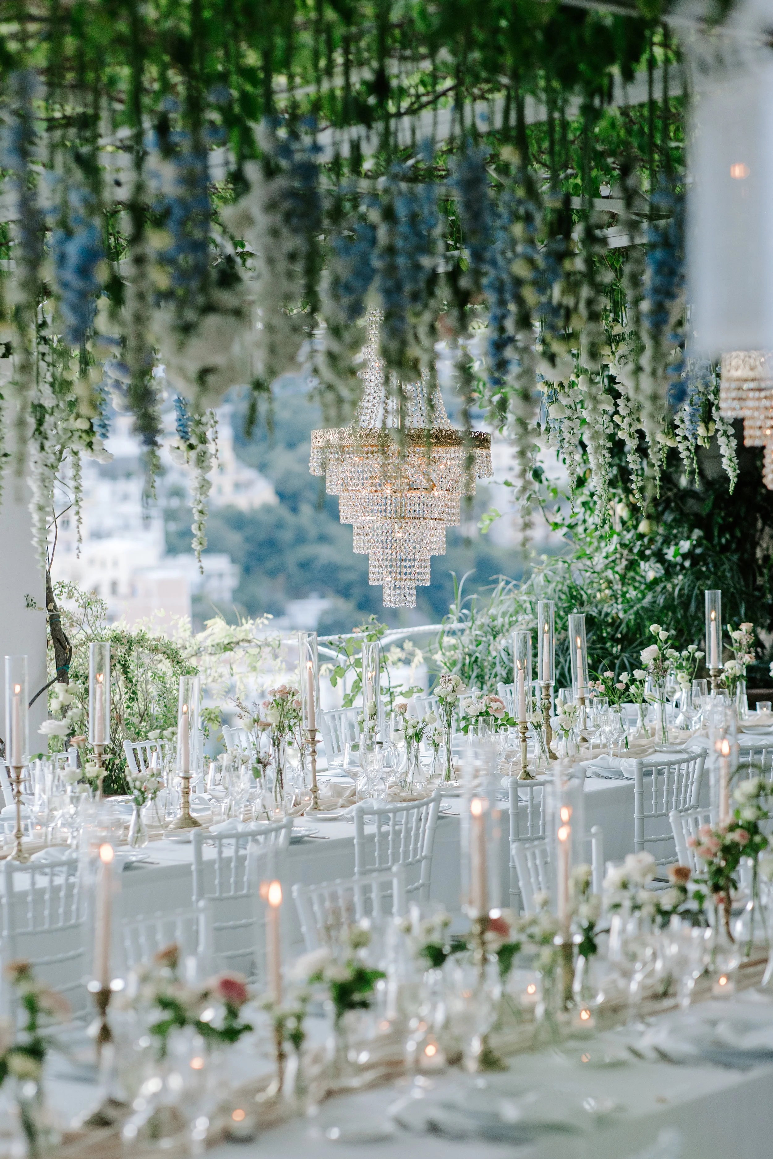Elegant indoor wedding reception with a long table set with white chairs, floral centerpieces, tall candles, and a chandelier hanging from above. Greenery and hanging flowers decorate the space with a scenic view in the background.