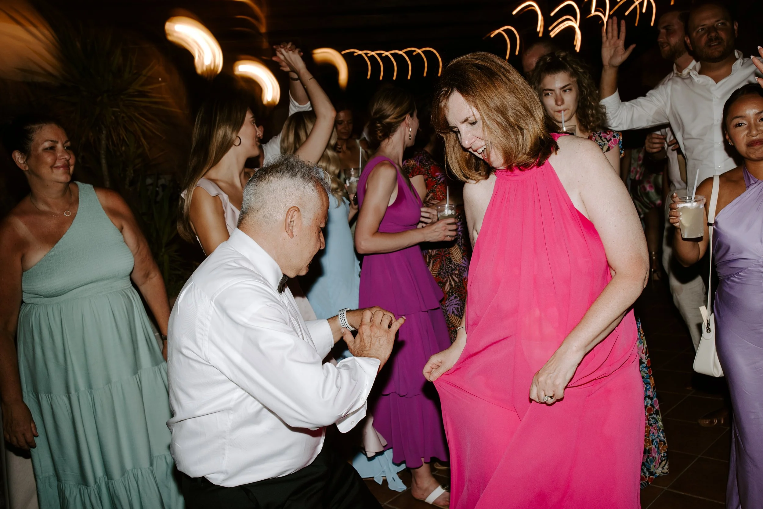 A woman in a pink dress is dancing at a party while a man kneels in front of her, holding her hand. Others in colorful dresses surround them, some holding drinks, and people are dancing and enjoying the celebration.