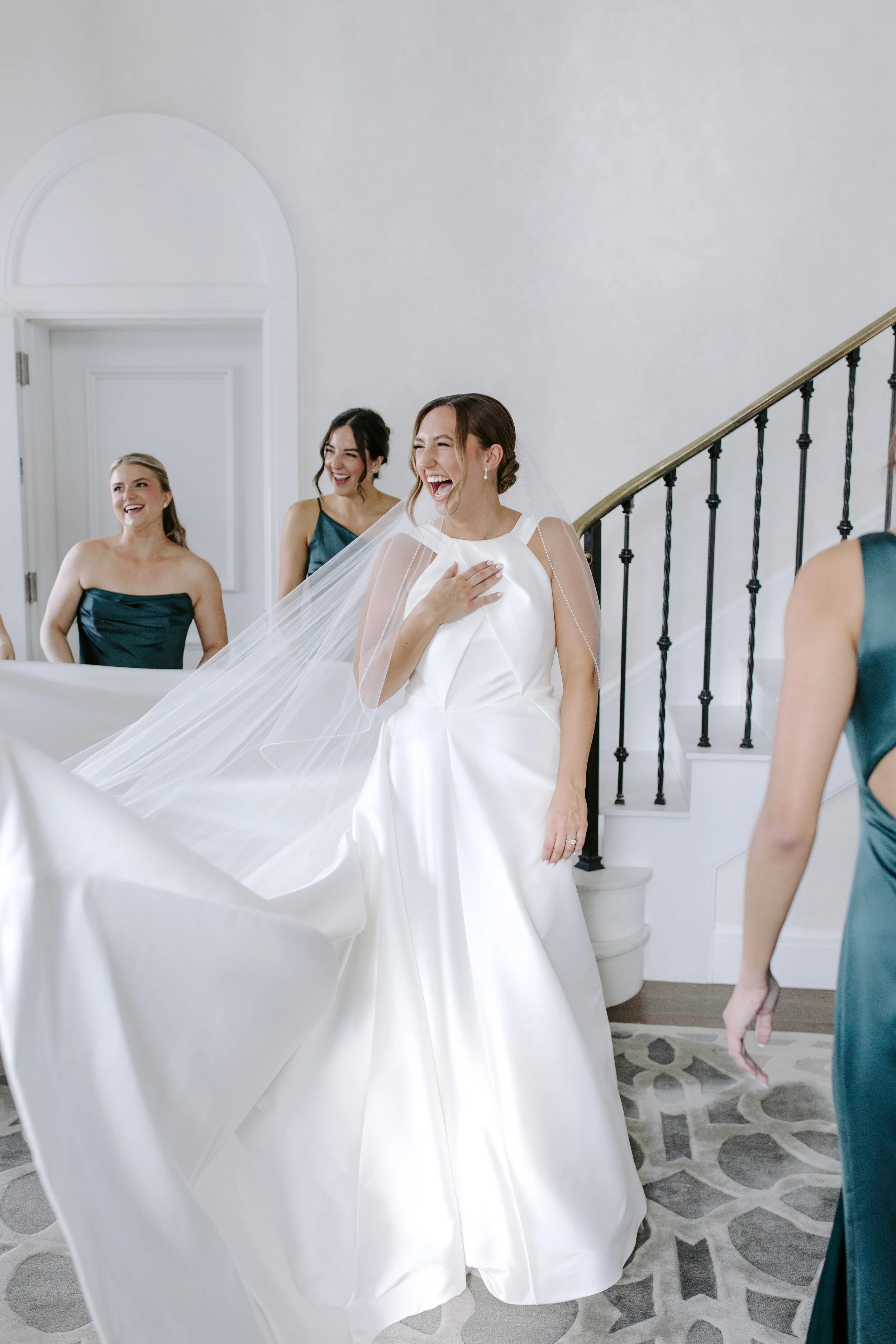 A bride in a white wedding gown and veil, laughing and touching her chest, surrounded by smiling bridesmaids in teal dresses indoors near a staircase.