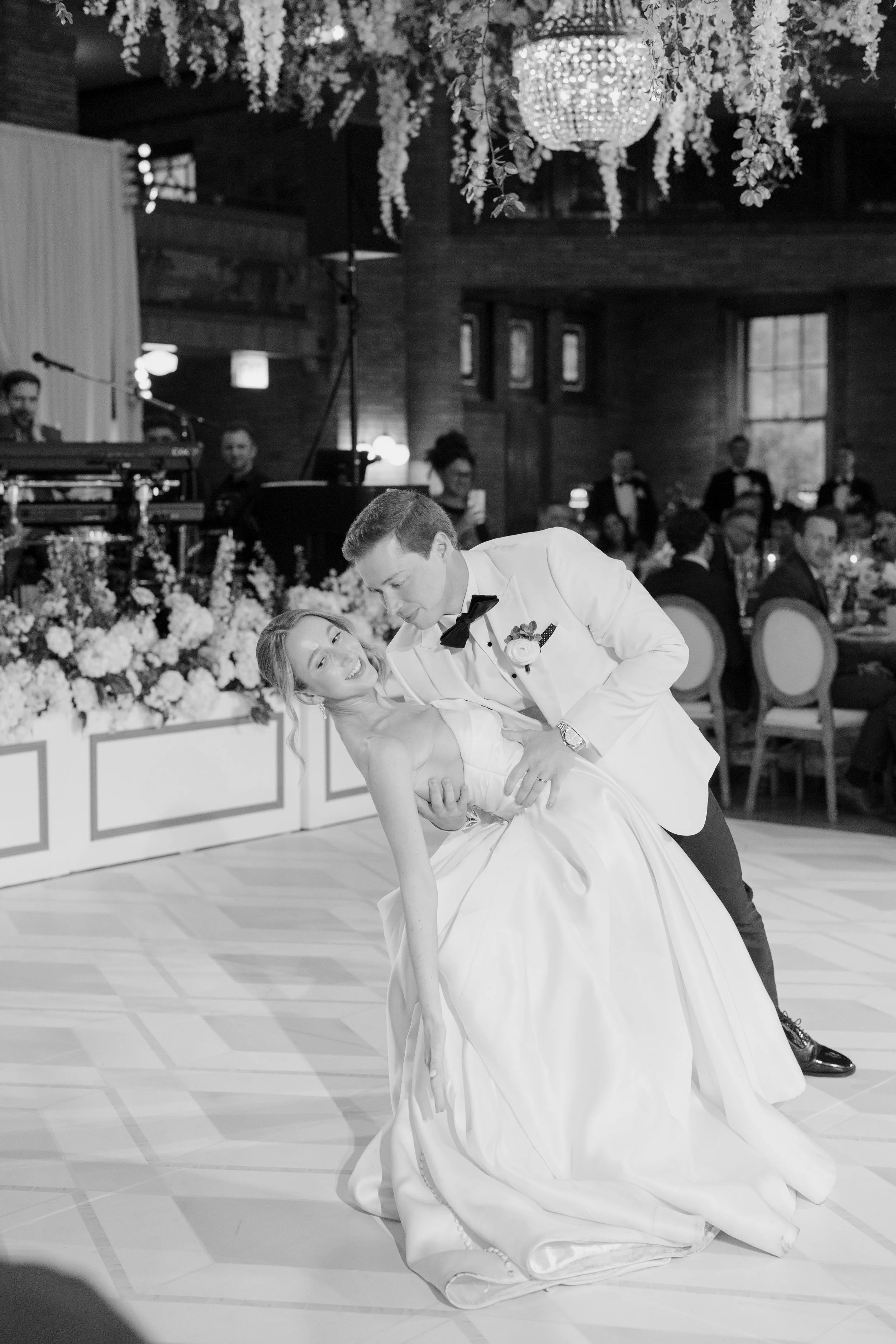 A black and white photo of a wedding reception with a bride and groom dancing. The bride is in a white wedding gown and the groom is in a tuxedo with a bow tie. Others are seated at tables in the background, and floral arrangements adorn the room.