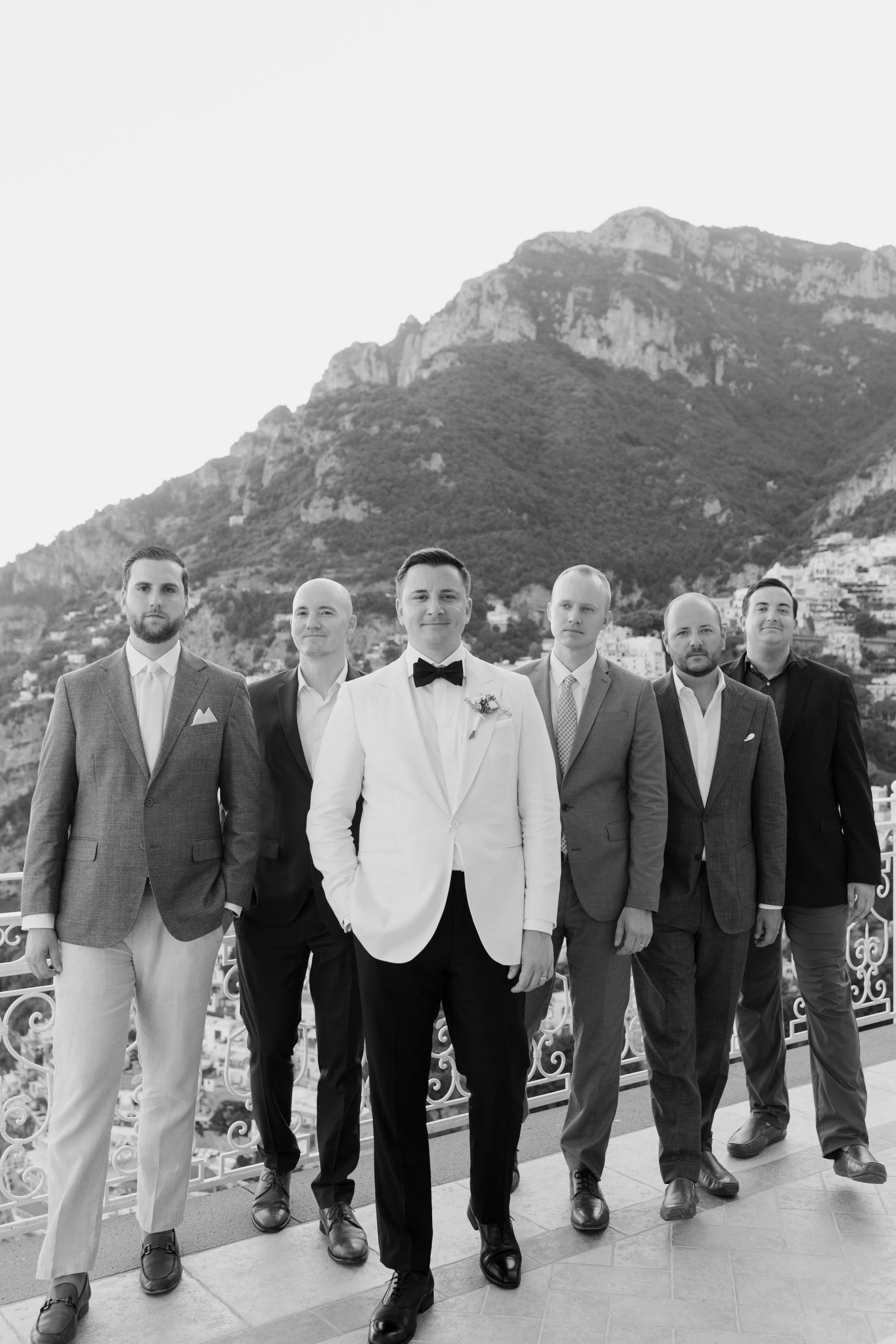 A group of six men dressed in formal suits standing on a balcony with a mountain in the background. The men are smiling and standing close together, with the central man wearing a white tuxedo with a black bow tie. The photo is in black and white.