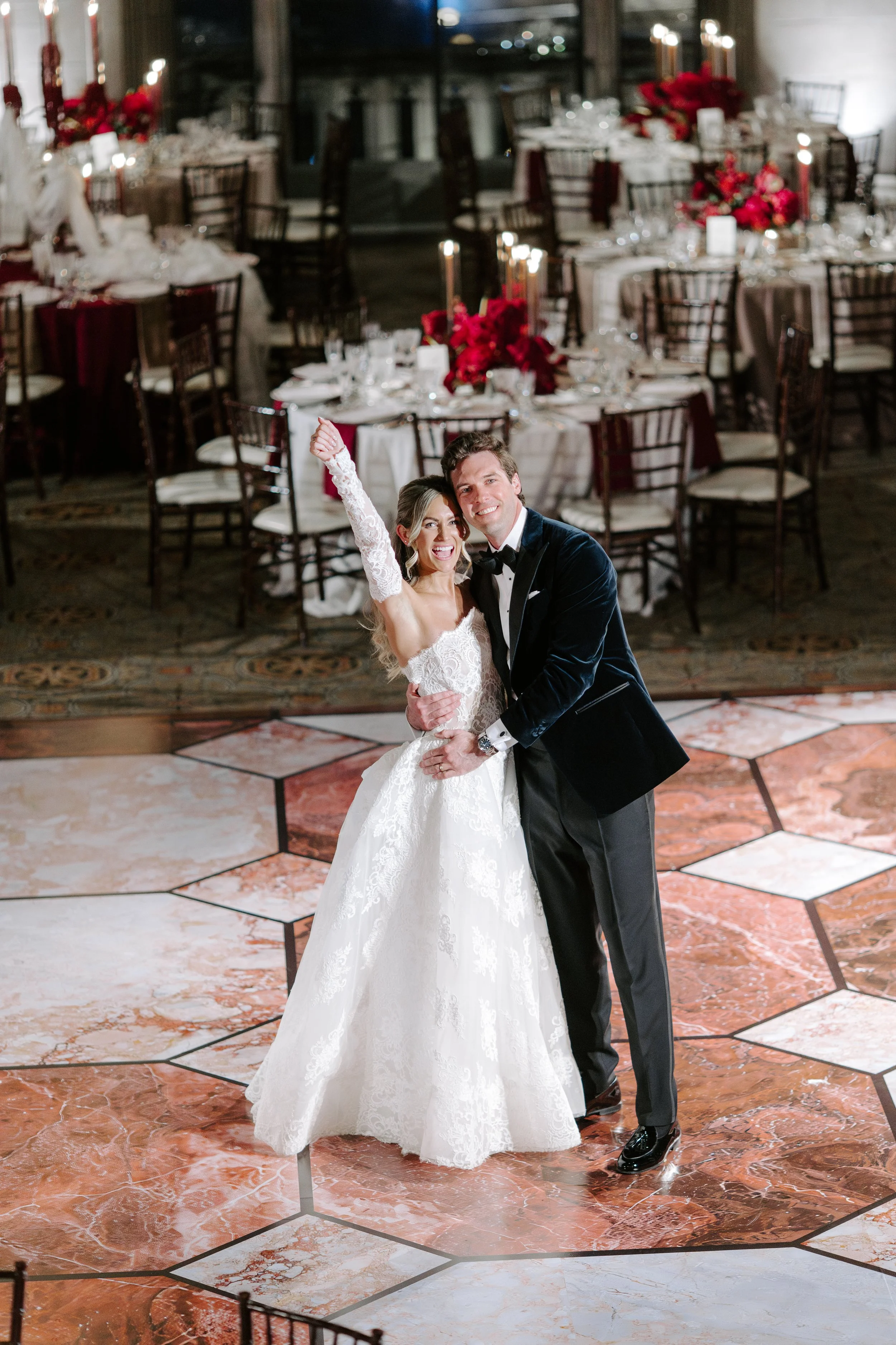 A bride and groom dancing at their wedding reception. The bride is wearing a white lace wedding gown and the groom is in a black tuxedo with a bow tie. The background features decorated tables with red floral centerpieces and candlelit candles.