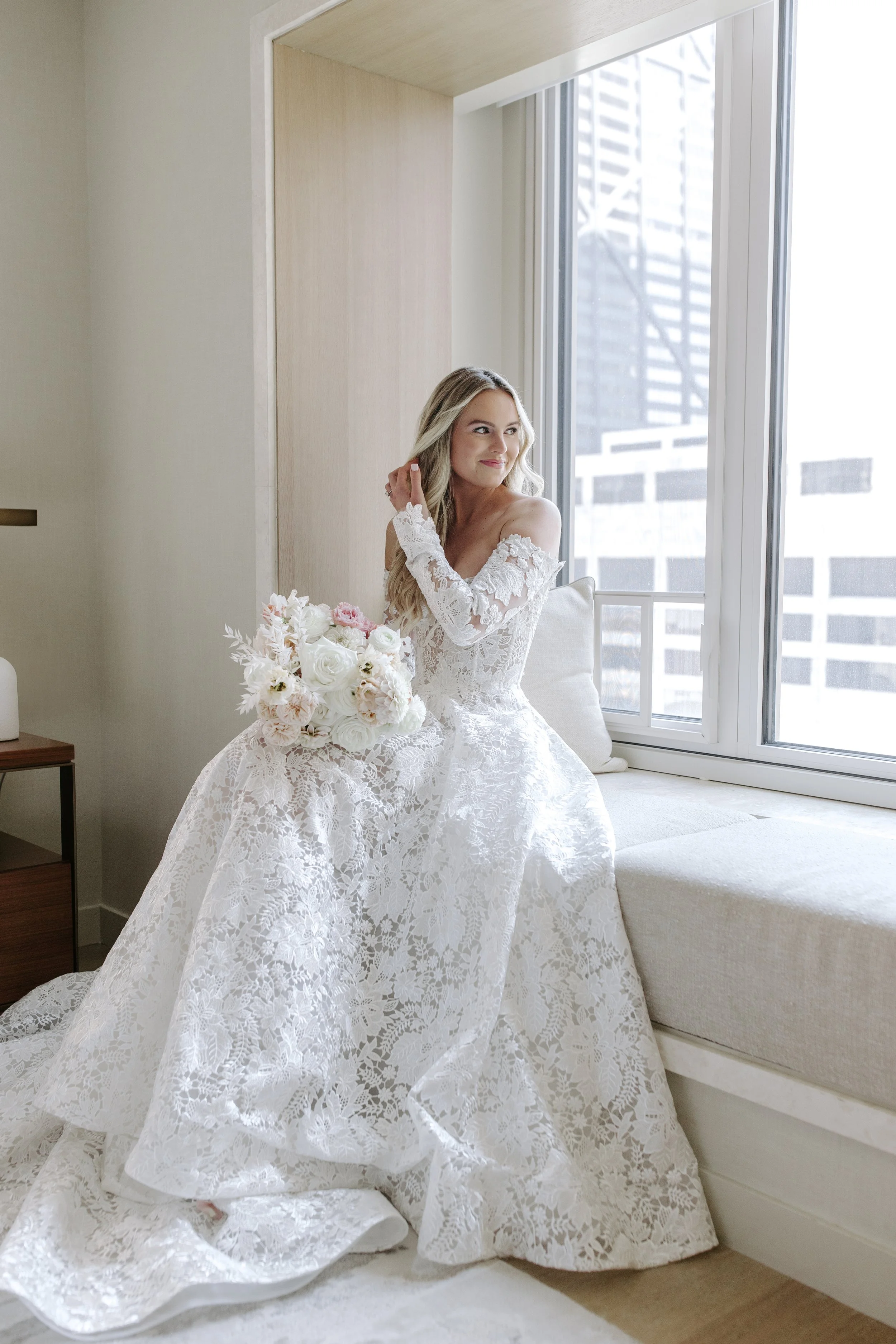 A woman dressed in a white lace wedding gown sitting on a window seat, holding a bouquet of flowers, and looking out the window with a gentle smile.