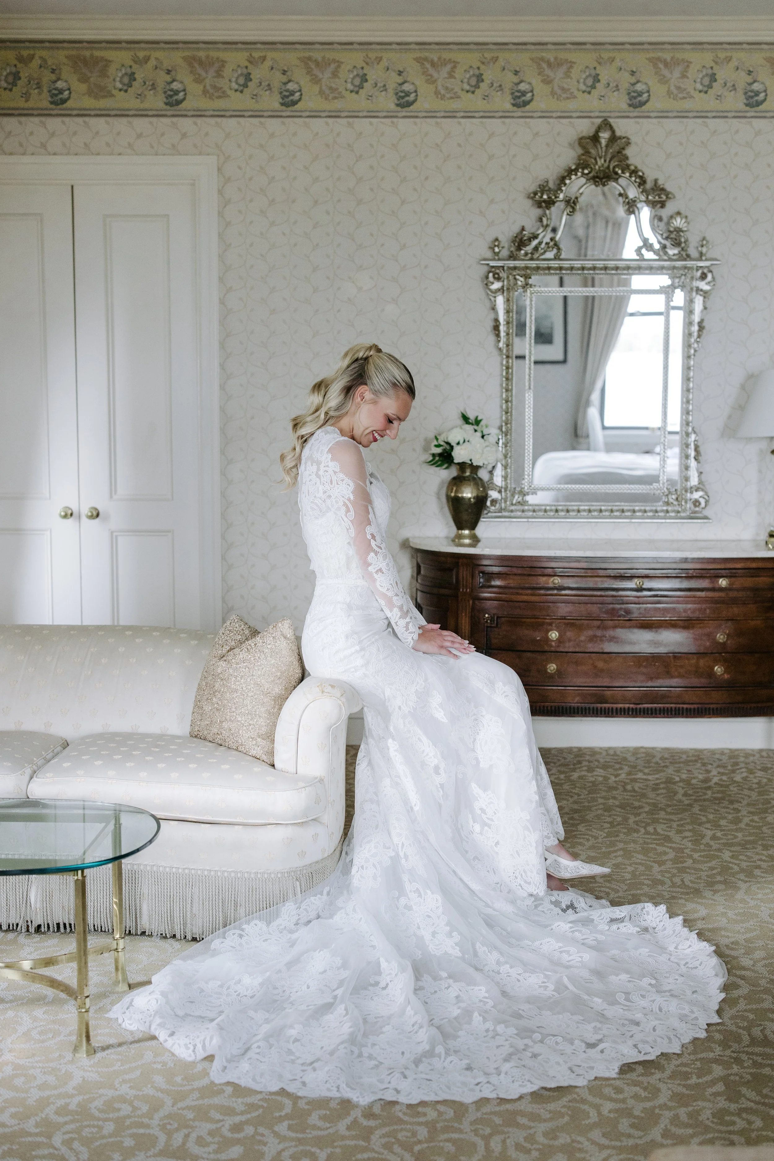 A woman in a white wedding dress sitting on a white sofa, smiling and looking down, with her train extended on the carpeted floor, in a well-decorated room with a large ornate mirror, a wooden dresser, a vase of white flowers, and a window with curta