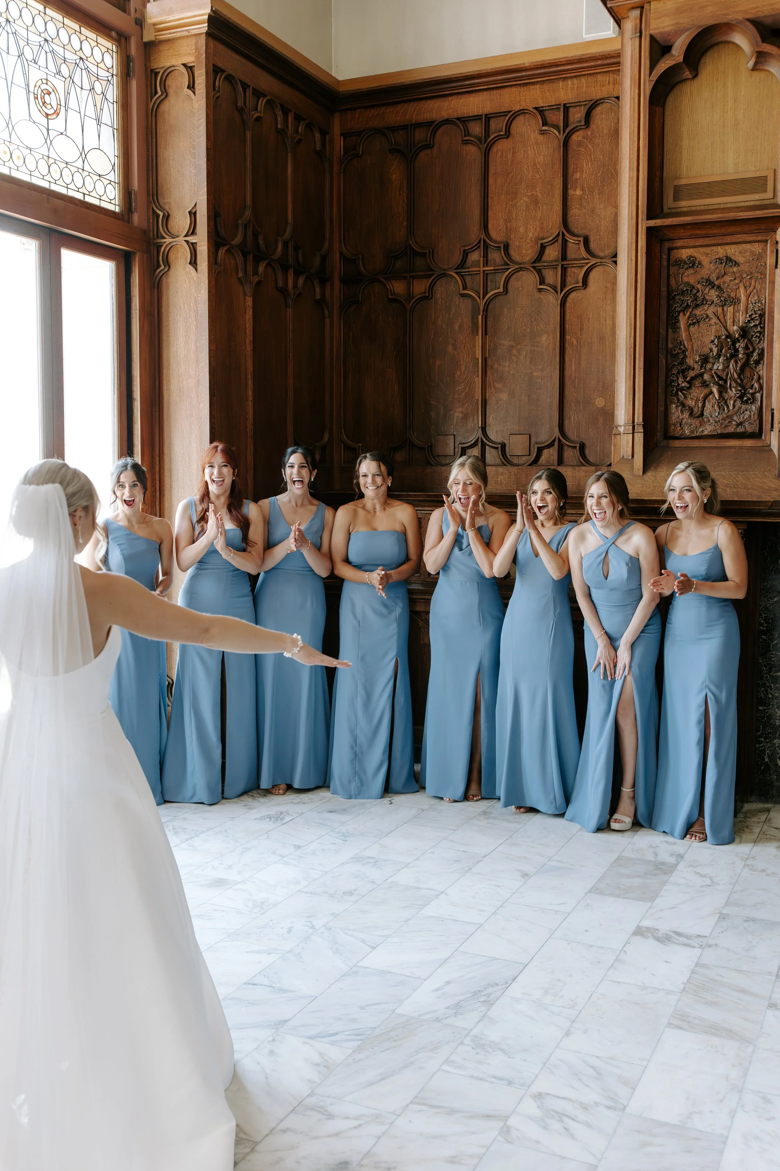 A bride in a white wedding dress with veil facing a group of eight women in matching blue dresses, celebrating indoors with wooden wall paneling and a marble floor.