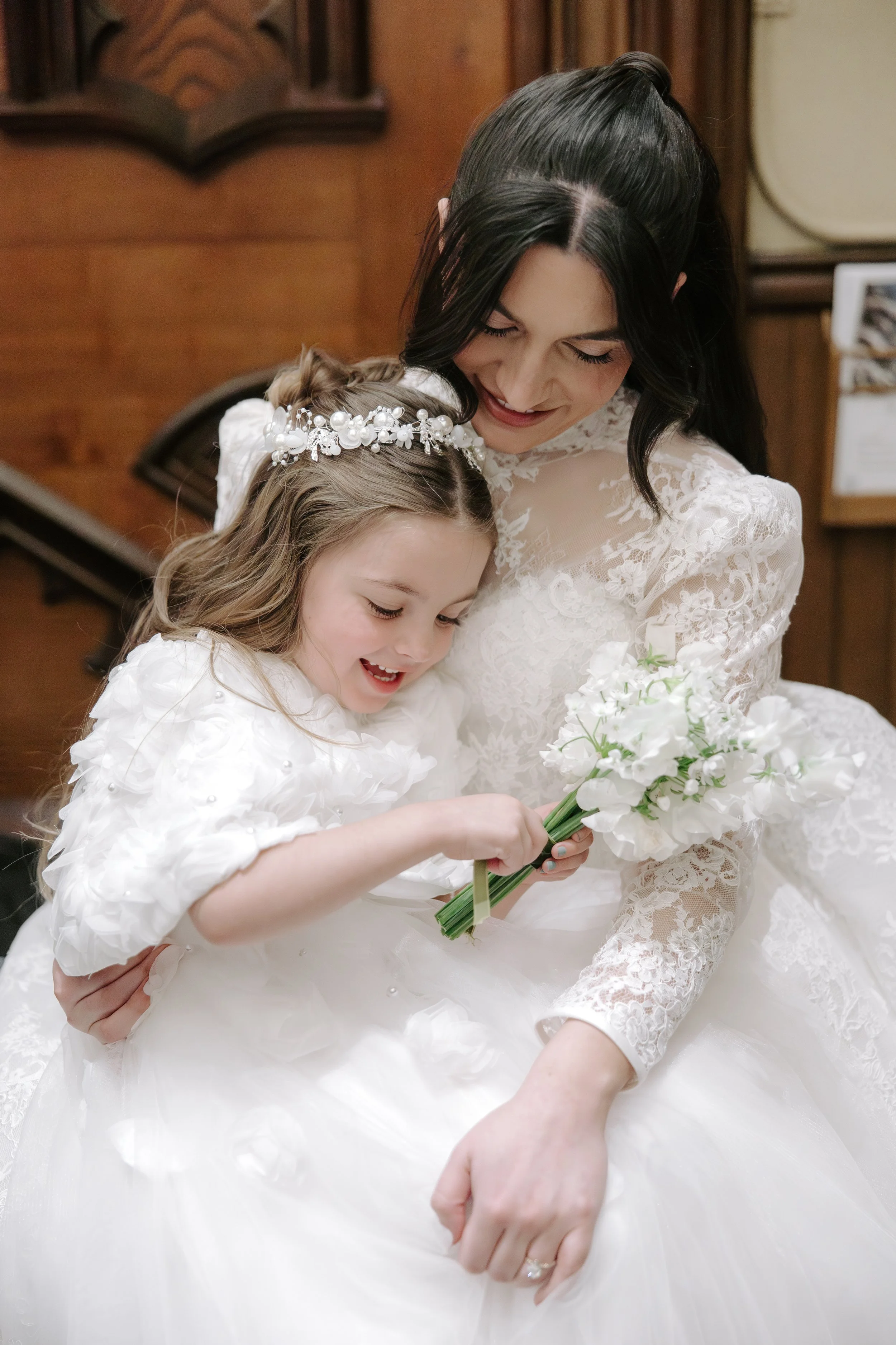 A woman in a lace wedding dress hugging a young girl in a white dress with a pearl and floral headband, both holding a bouquet of white flowers.