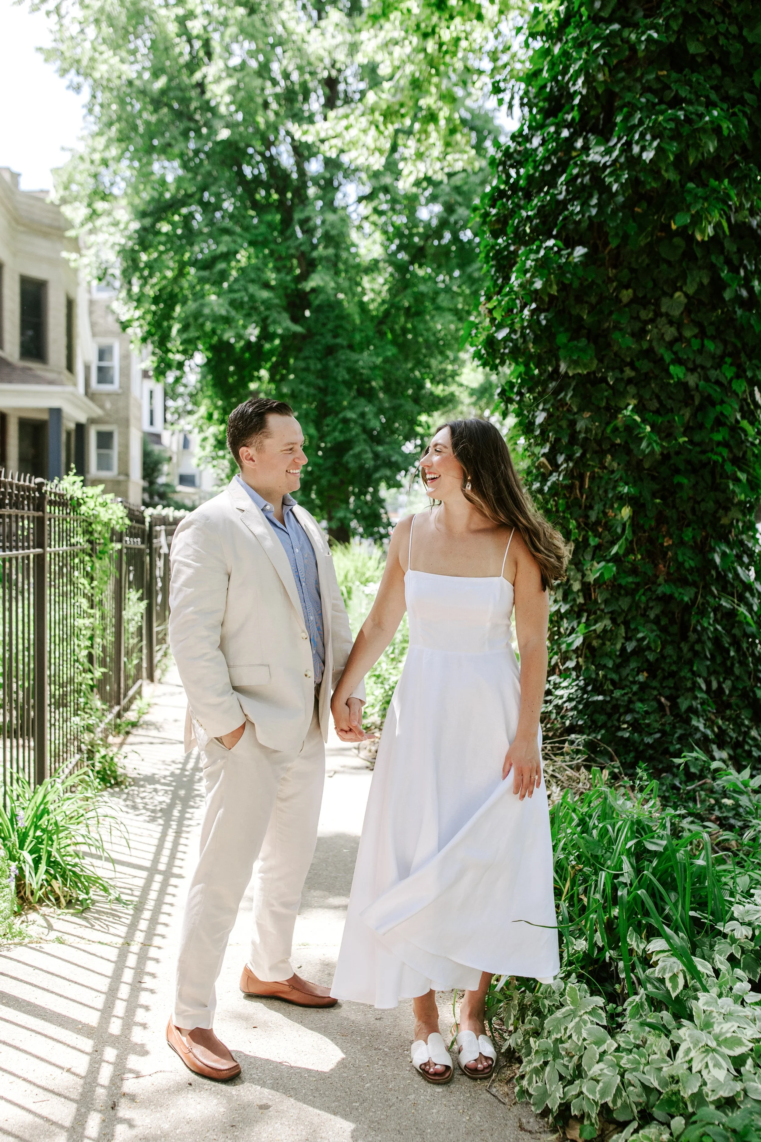 A couple holding hands and smiling during an outdoor wedding ceremony on a sunny day. The groom is wearing a cream-colored suit with a light blue shirt, and the bride is in a white dress with spaghetti straps. They are standing on a sidewalk with lus