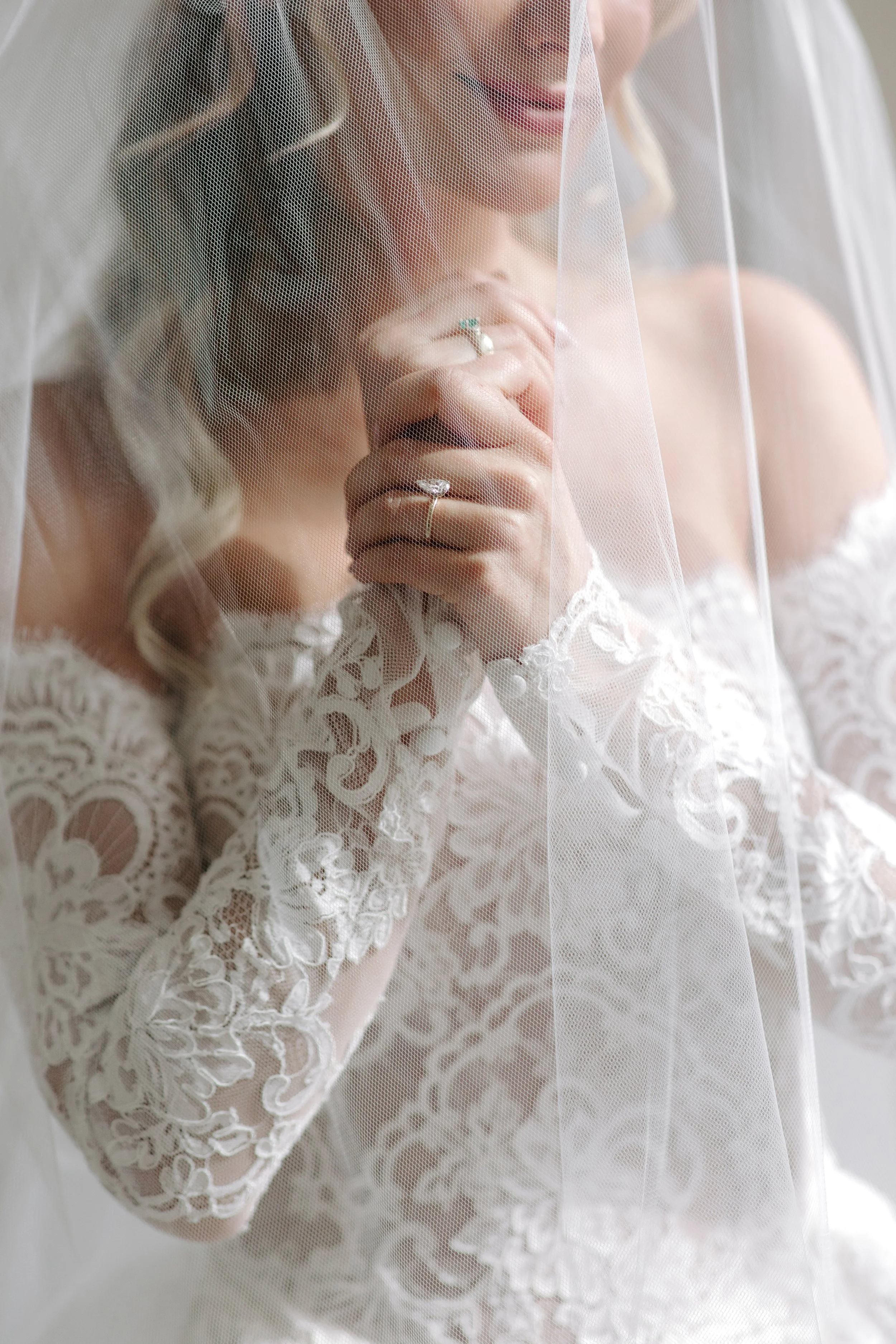 Close-up of a woman wearing a lace wedding dress and veil, with her hand near her face, showing rings on her fingers.