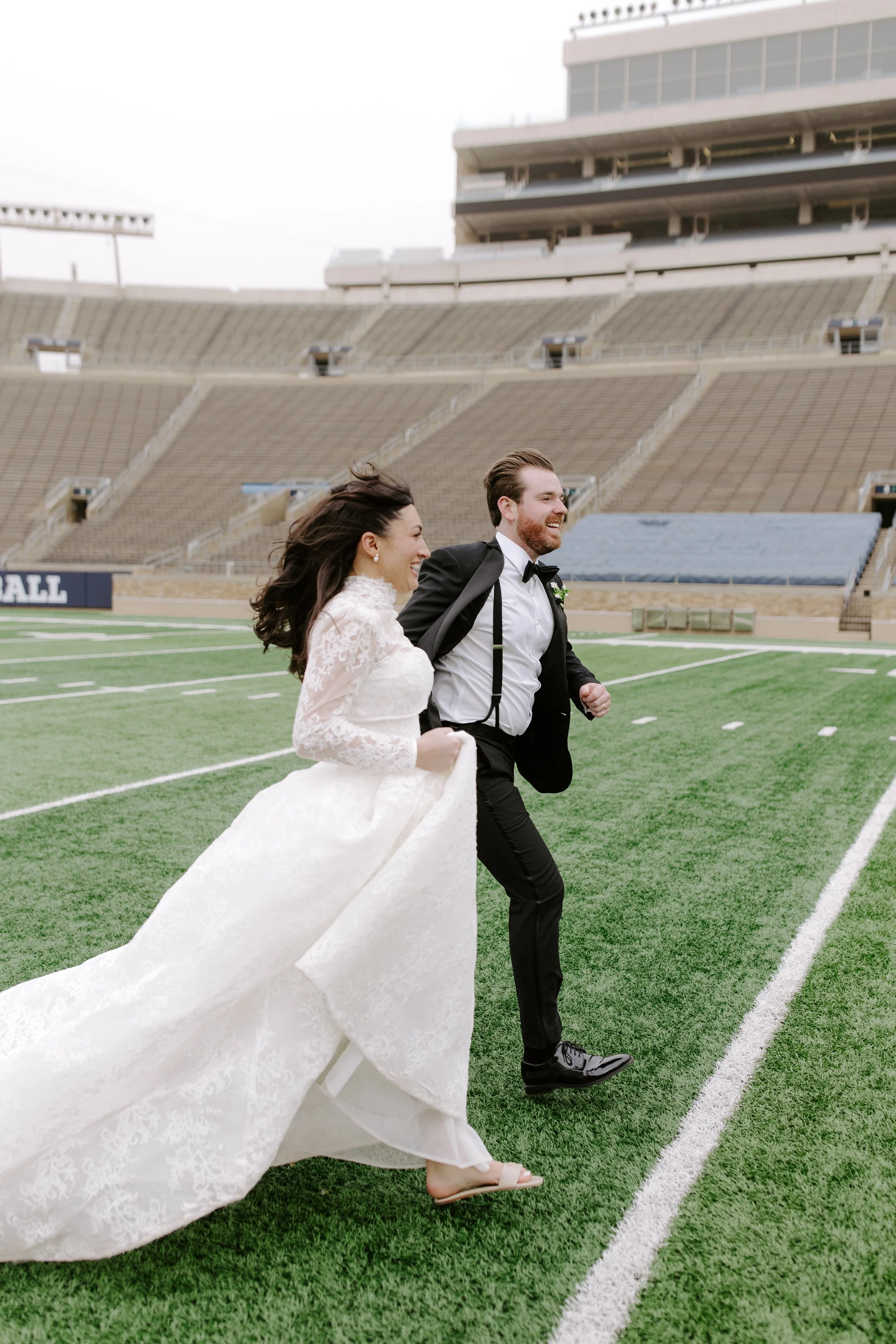 A bride and groom running on a football field at a stadium, dressed in wedding attire, smiling and laughing.