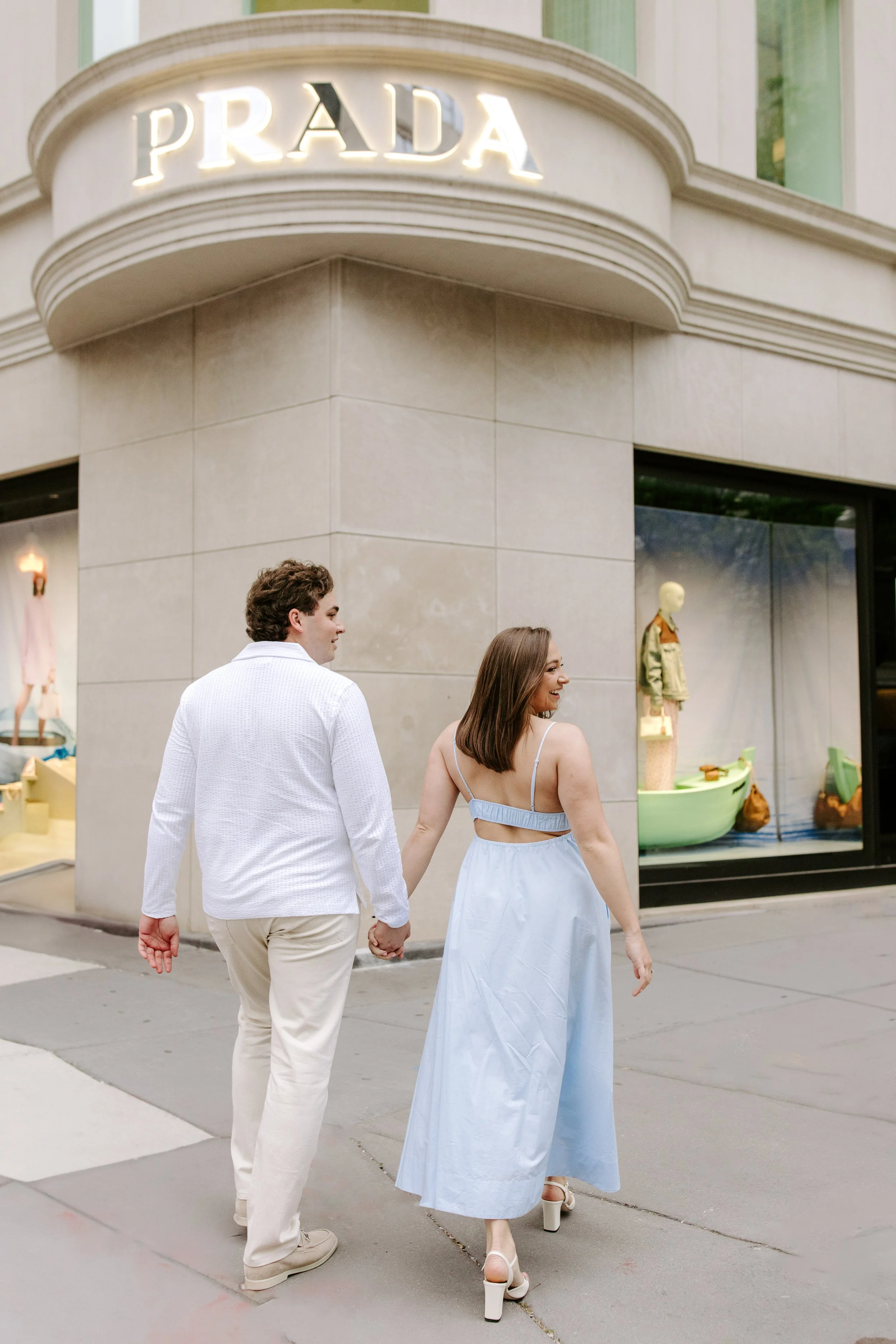 A young couple walking hand in hand on a city sidewalk, holding hands and smiling. The woman wears a light blue flowy dress and high-heeled shoes, while the man wears a white shirt and beige pants. They are in front of a Prada store with mannequins d