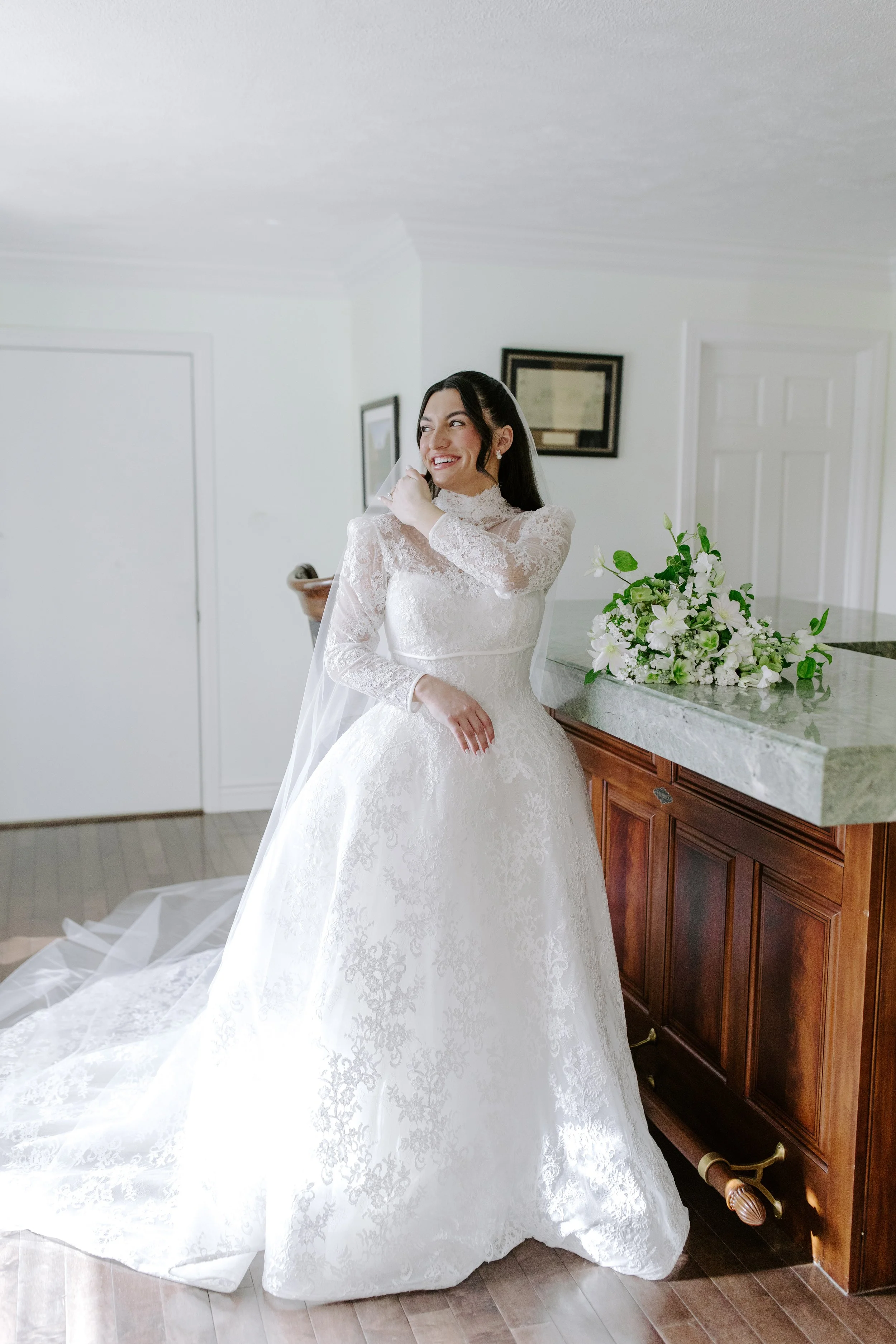 A bride in a white lace wedding gown with long sleeves, standing indoors near a wooden dresser with a bouquet of white flowers on top, smiling and adjusting her veil.