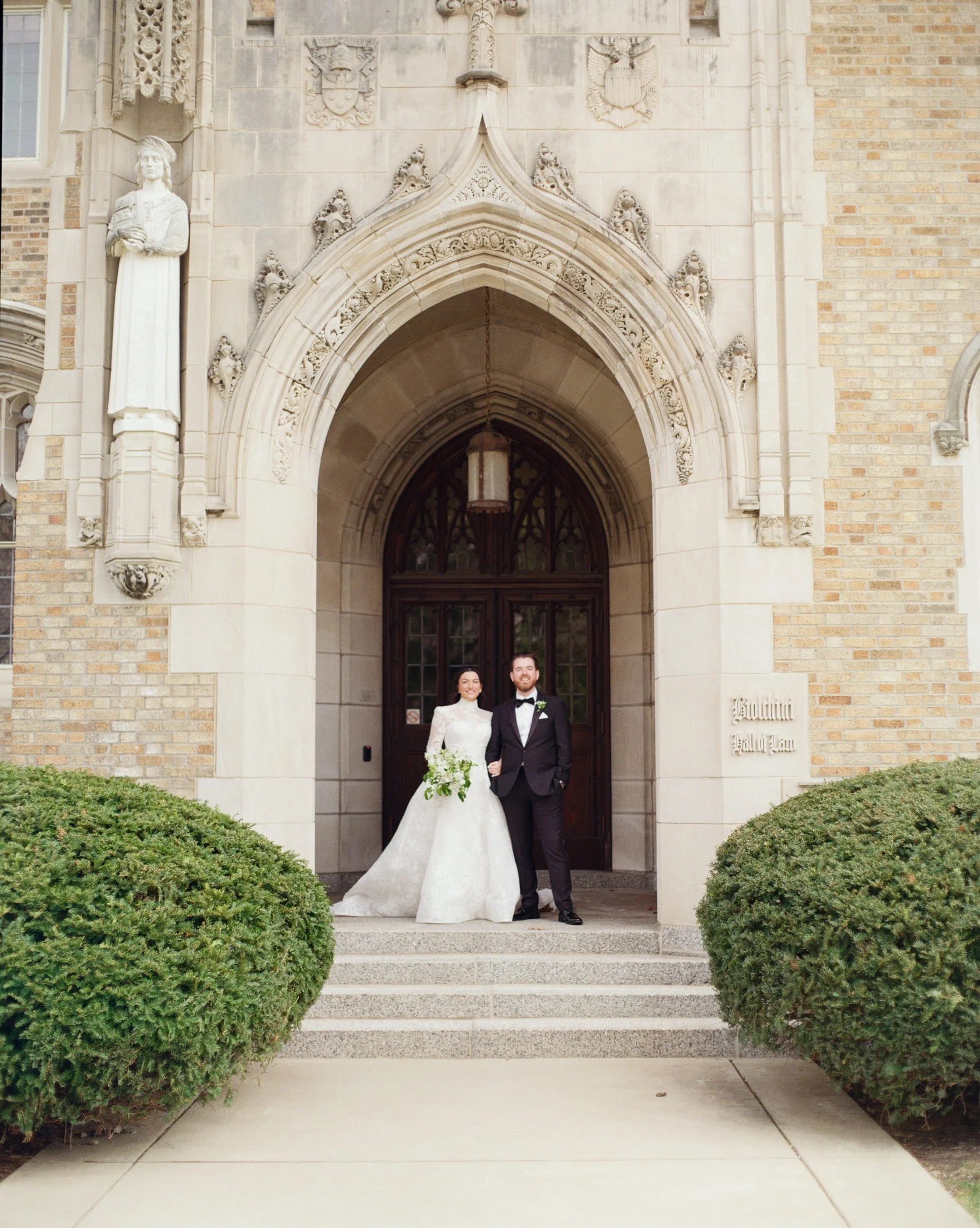 A newlywed bride and groom standing on the steps of a church, smiling, with green bushes on either side of the stairs. The church has an arched doorway, stone and brick facade, and sculptures above the entrance.