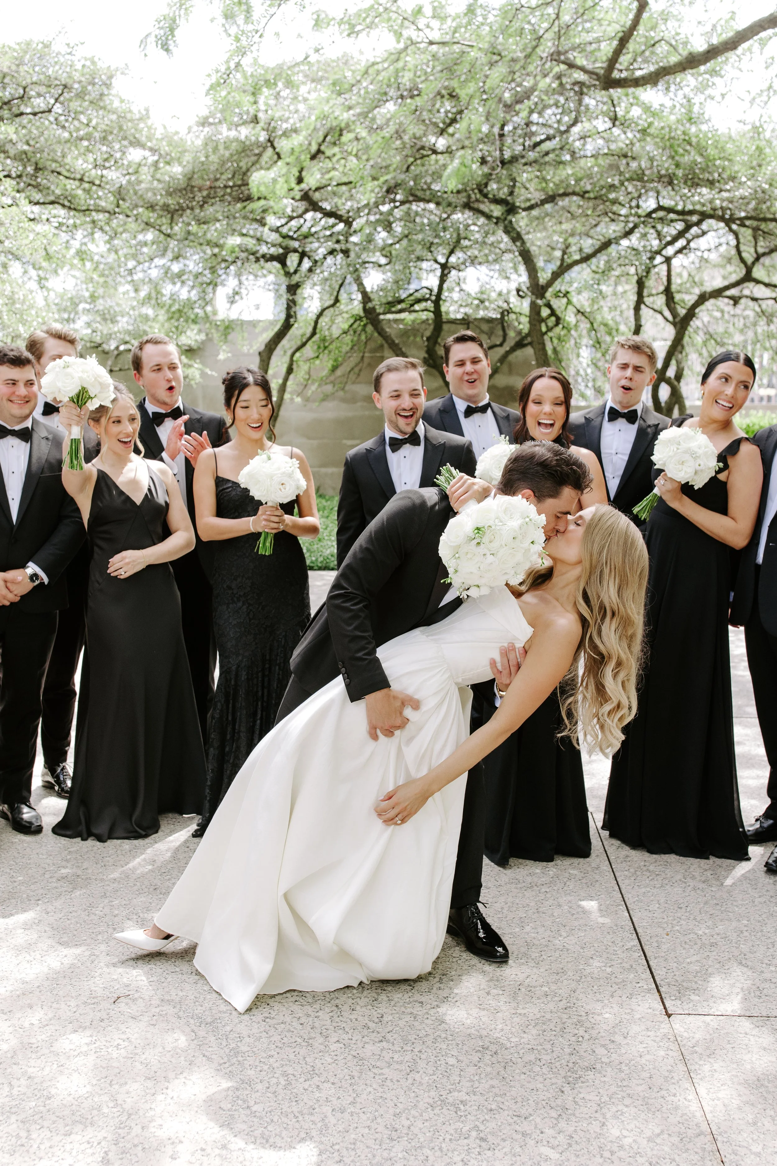 A bride and groom share a kiss while the wedding party looks on and laughs under a large tree with blooming branches.