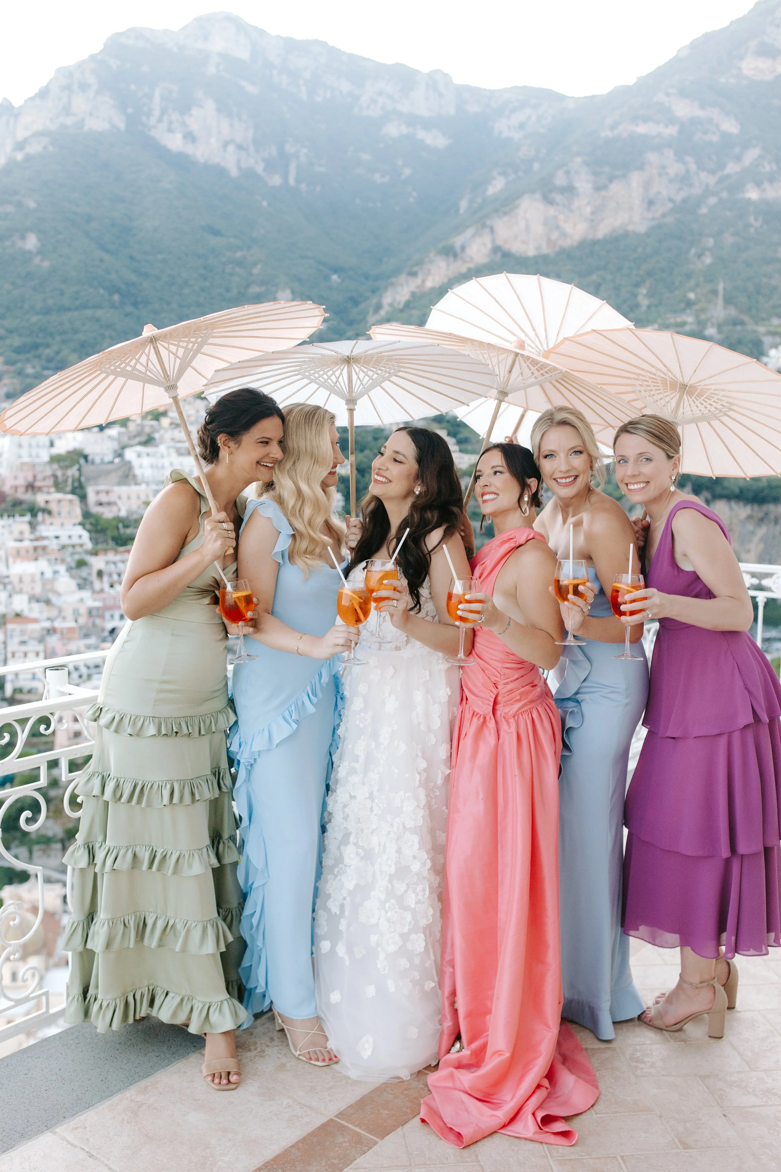 A group of seven women in colorful dresses holding orange drinks with umbrellas, standing on a balcony with a scenic mountainous backdrop.